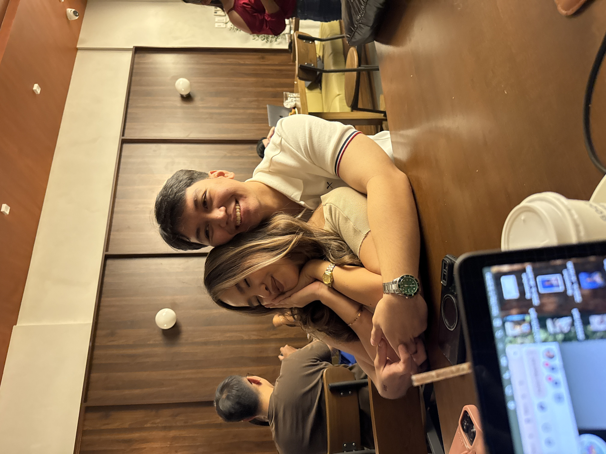 A young couple is seated at a dark brown table indoors. The man, with short black hair and a white polo shirt, is smiling while leaning on the woman. The woman, with long brown hair and a light-colored top, is resting her head on her hands. Both are wearing watches. A laptop, a cup, and a pink phone are also on the table. In the background, there are other people seated, and the walls are paneled with horizontal dark brown wood. Based on the interior and ambiance, the location might be a coffee shop in Davao City, Philippines.