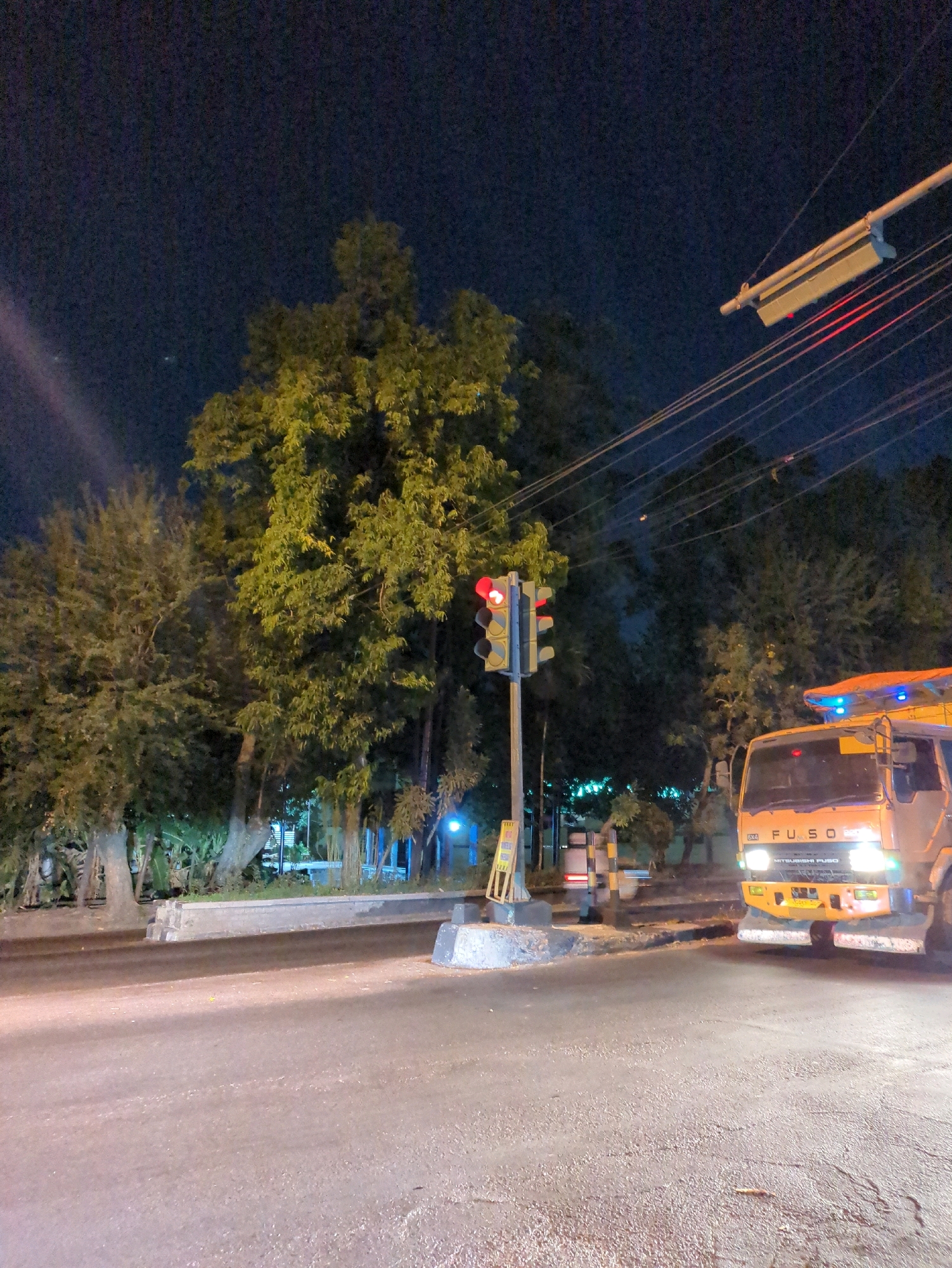 An orange Fuso truck is parked on the side of a wet road at night, illuminated by its headlights. A traffic light displays a red signal. Tall trees with dense foliage line the background, and electrical wires crisscross overhead. The scene appears to be a roadside in Surakarta, Indonesia, possibly near a wooded area.