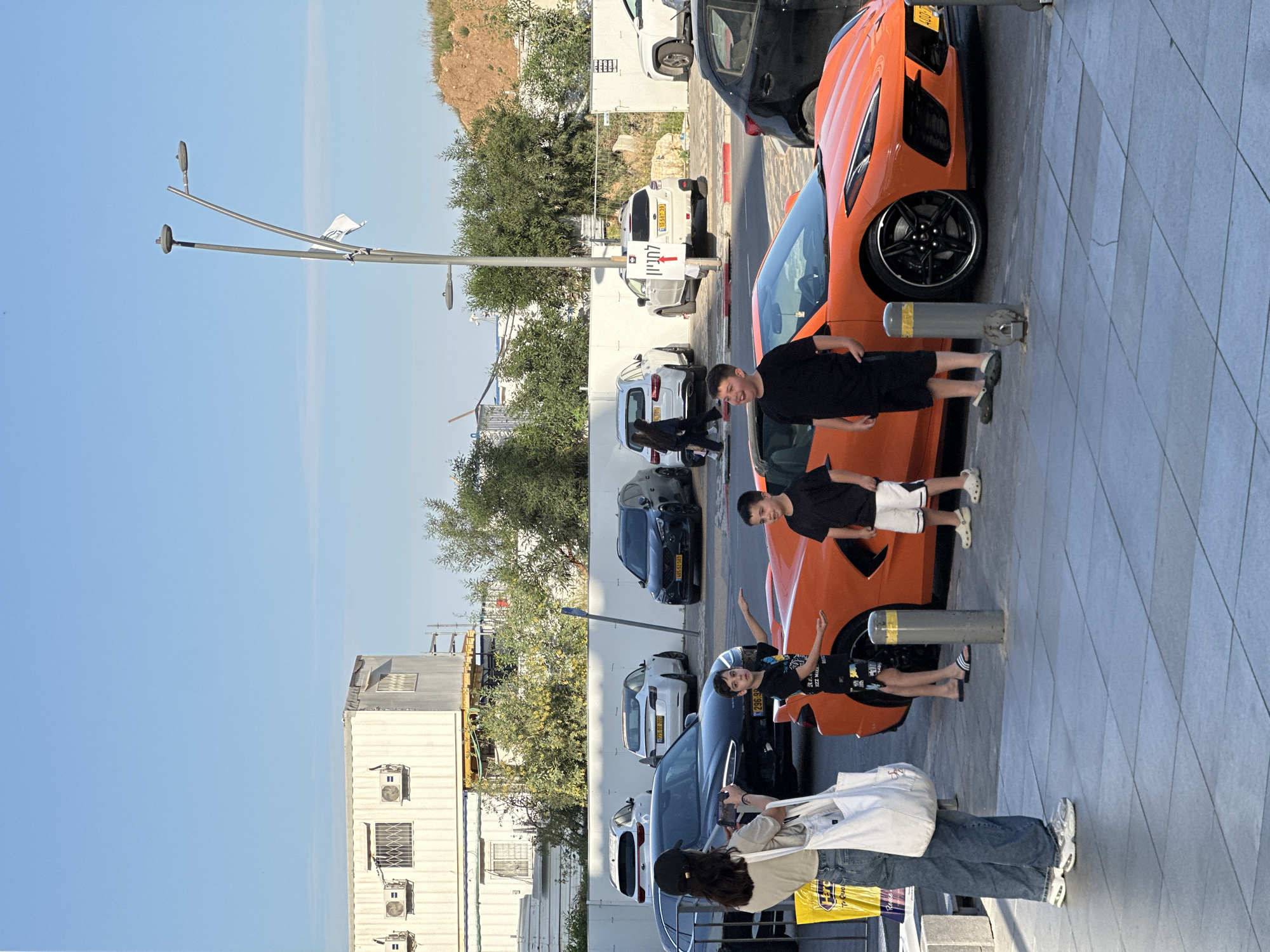 The image shows a parking area in Herzliya, Israel, with multiple parked cars and several people standing on the sidewalk. The sky is clear and blue. A tall street light stands on the left.
A bright orange sports car is parked along the edge of the sidewalk. Three boys appear to be posing or leaning against the car near its wheels. An adult female with a white bag stands on the sidewalk.