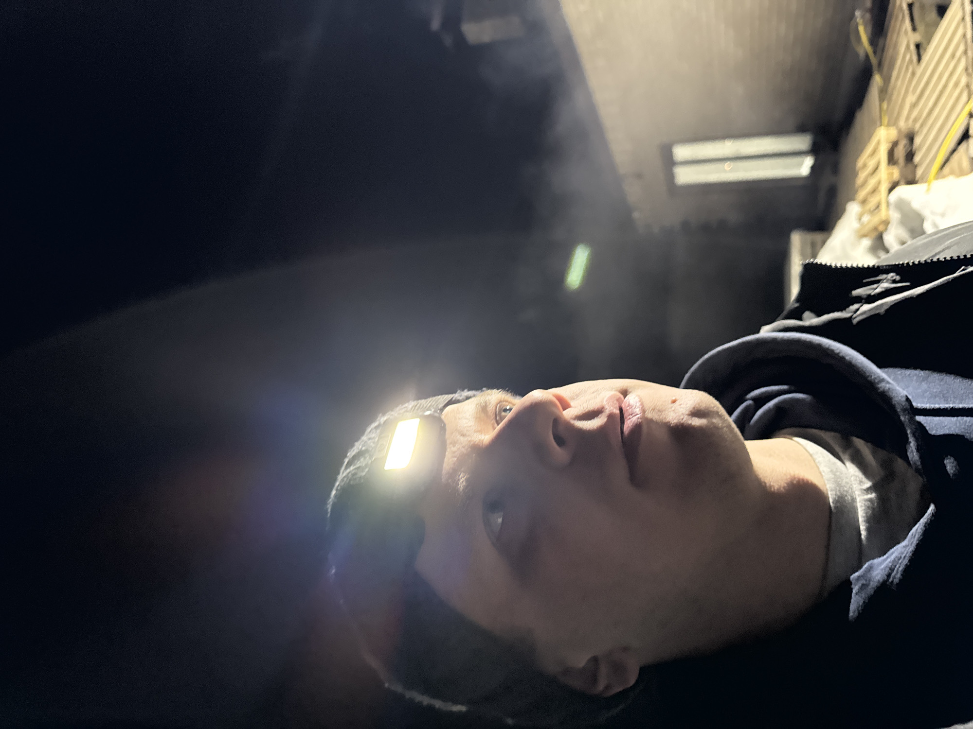 A low-angle shot shows a person lying down, illuminated by a headlamp. The location appears to be an enclosed space, possibly a warehouse or industrial setting, given the presence of a pallet stack and fluorescent lighting in the background. The person is wearing a beanie and a hooded jacket, looking upwards. The environment appears dark and somewhat damp, as indicated by the presence of mist or vapor in the air. The location is likely Gornyi (Dvurechenskoe S/P), Russia, based on the prompt.