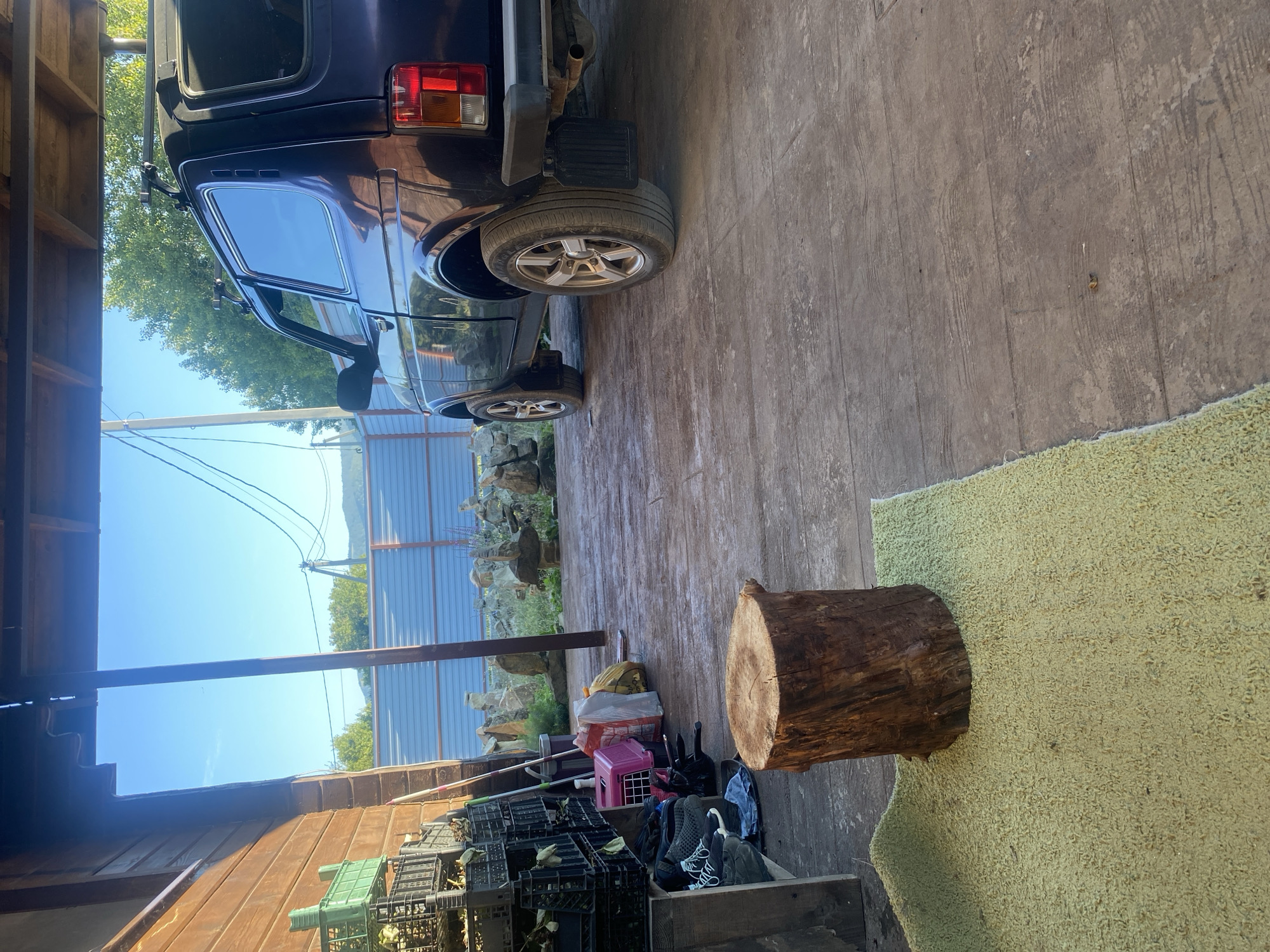 The image shows an outdoor area, possibly a yard or a covered porch, in the village of Aderbievka, Russia. In the foreground, a light green rug is placed on a wooden floor, and on top of the rug is a short, thick tree stump. To the right of the stump is a dark-colored SUV parked on the wooden floor. The vehicle appears to be a Niva. Behind the SUV, a blue metal fence can be seen, with some rocks and vegetation visible beyond it. To the left of the SUV, stacked plastic crates are visible, some containing what appear to be plants or produce. A pink pet carrier, bags, and shoes are also present near the crates. The background shows trees, a clear blue sky, and power lines, suggesting a rural or suburban setting. The lighting indicates it is daytime, likely morning or afternoon, with the sun casting shadows. There are no people visible in the image.