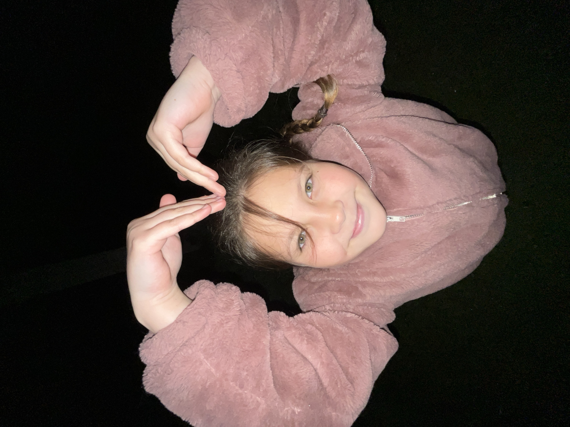 A young girl is posing for a photo. She is wearing a pink, fluffy jacket and has light brown hair with a braid visible. She is smiling and making a hand gesture with both hands. The background is dark, suggesting it might be nighttime. The location is not discernible from the image. There is no information to suggest the location is Novopokrovskaya, Russia.