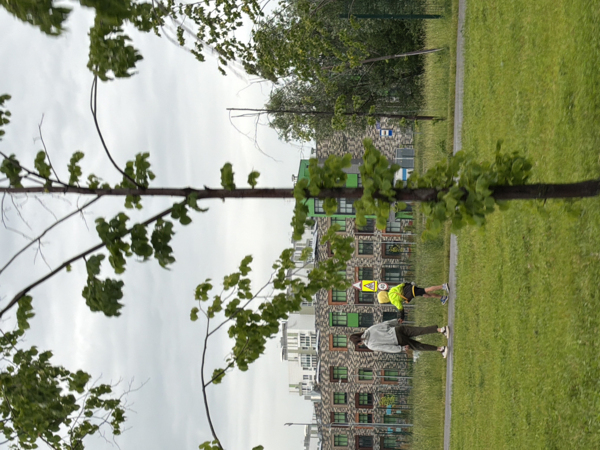 This image captures two individuals in an outdoor setting, with modern buildings in the background and a grassy field. One person, wearing a bright yellow jacket and a yellow cap, is bent over with their hands on the ground, possibly engaged in an exercise or stretching routine. They are also wearing dark shorts and athletic shoes. The other person, dressed in a grey long-sleeved shirt and dark pants, stands upright and appears to be walking or standing on a paved path. They are wearing sneakers.
The scene is characterized by lush green grass and trees with vibrant green leaves, suggesting it is daytime, likely spring or summer. The sky is overcast, indicating a cloudy or possibly pre-rainy weather condition. The buildings in the background are multi-story structures with a distinctive architectural style, featuring a patterned facade and numerous windows, some of which are accented with green frames. A road sign with a yellow triangular warning symbol and the number "40" is visible, hinting at a speed limit or caution. Another blue and white sign is also present, though its text is not legible. The overall atmosphere appears casual and active, with the individuals likely enjoying outdoor recreation or exercise.