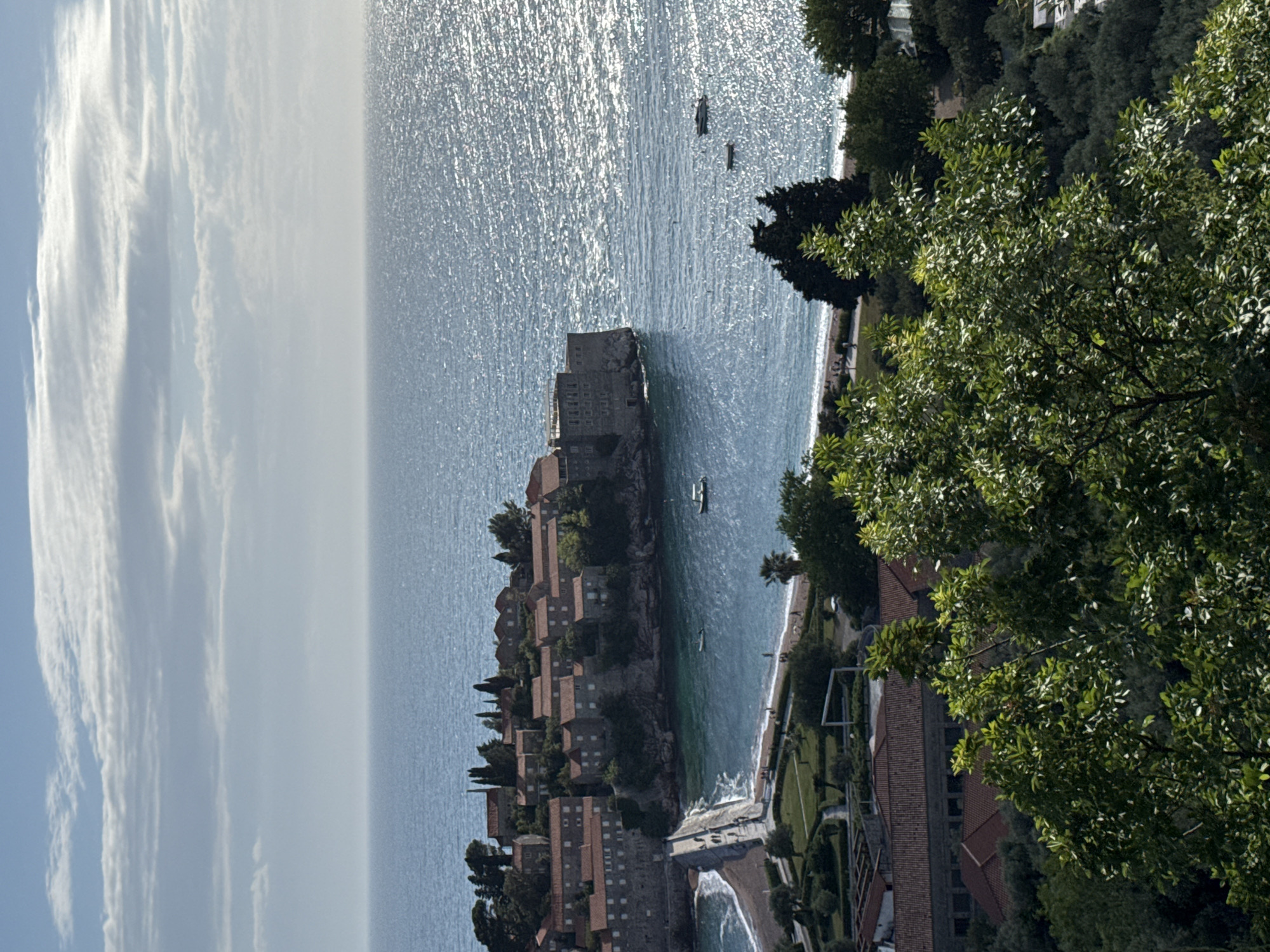 The image captures a high-angle, landscape view of Sveti Stefan, Montenegro, from an elevated position. In the foreground, a lush green tree fills the right side of the frame, partially obscuring the view. Below and to the left of the tree, sections of terra-cotta roofed buildings can be seen amidst greenery, suggesting a resort or hotel complex. The coastline extends from this area, leading to a peninsula topped with stone buildings resembling a medieval fortress or village. The peninsula is connected to the mainland by a narrow isthmus.
The Adriatic Sea dominates the central portion of the image, its surface shimmering in the sunlight. Small boats dot the water near the peninsula. The sky is bright with thin clouds on the left.