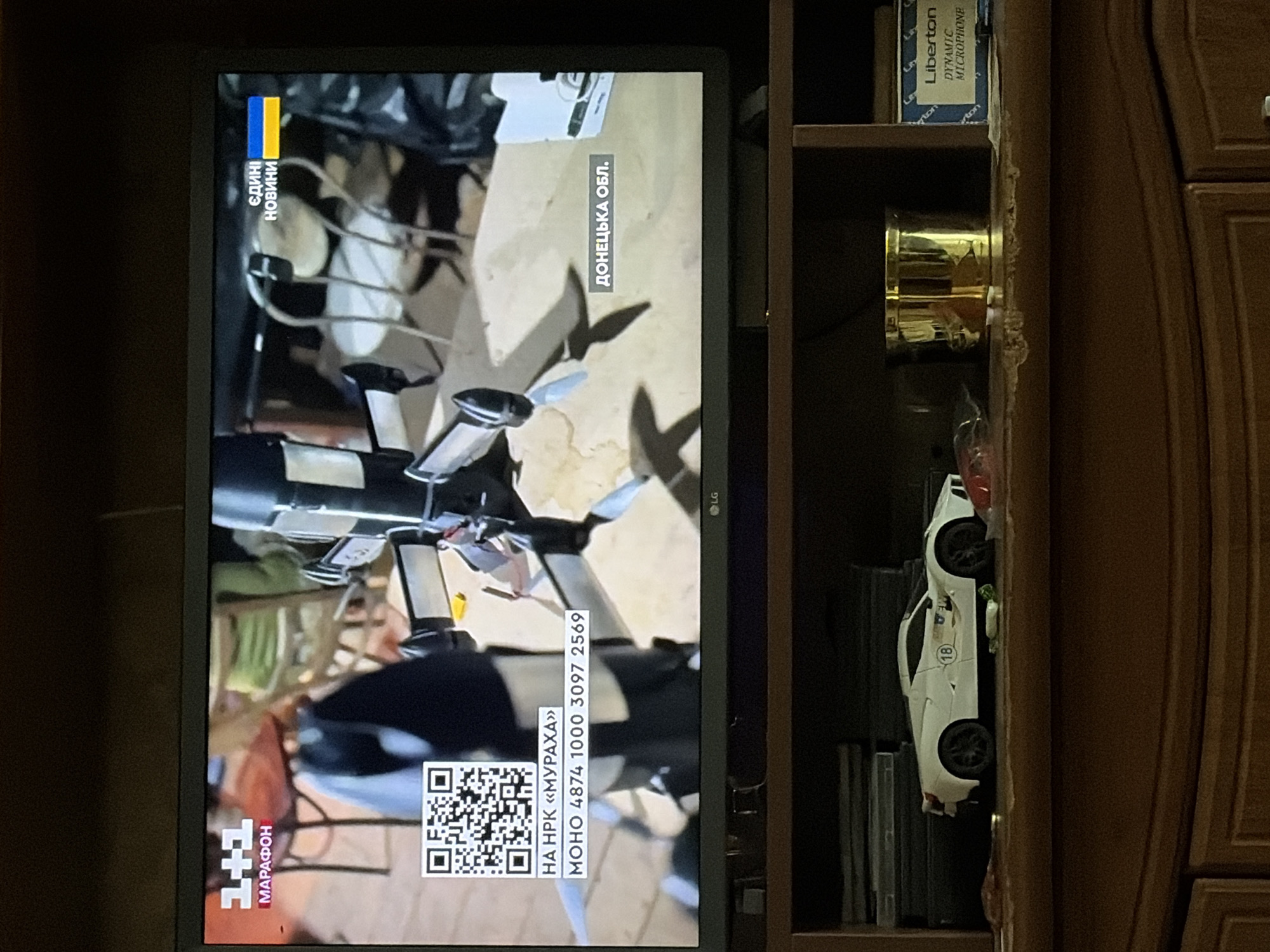 The image shows a television screen displaying a news broadcast from Ukraine. The screen displays a logo for "1+1 МАРАФОН" and a Ukrainian flag with the text "ЄДИНІ НОВИНИ" (United News). Below this, a QR code is visible, along with text in Ukrainian: "НА НРК «МУРАХА»" (On NPK "Muraha") and a phone number "МОНО 4874 1000 3097 2569". Another text overlay on the screen reads "ДОНЕЦЬКА ОБЛ." (Donetsk Oblast). The background of the broadcast appears to be an indoor scene with furniture and objects that are blurry.
Below the television, on a shelf, a white toy sports car is visible, along with a golden cylindrical object, a box with the brand name "Liberton" and the text "DYNAMIC MICROPHONE" printed on it, and some other items. The shelf appears to be made of wood and shows signs of wear and tear. The overall environment appears to be a home or an office setting.