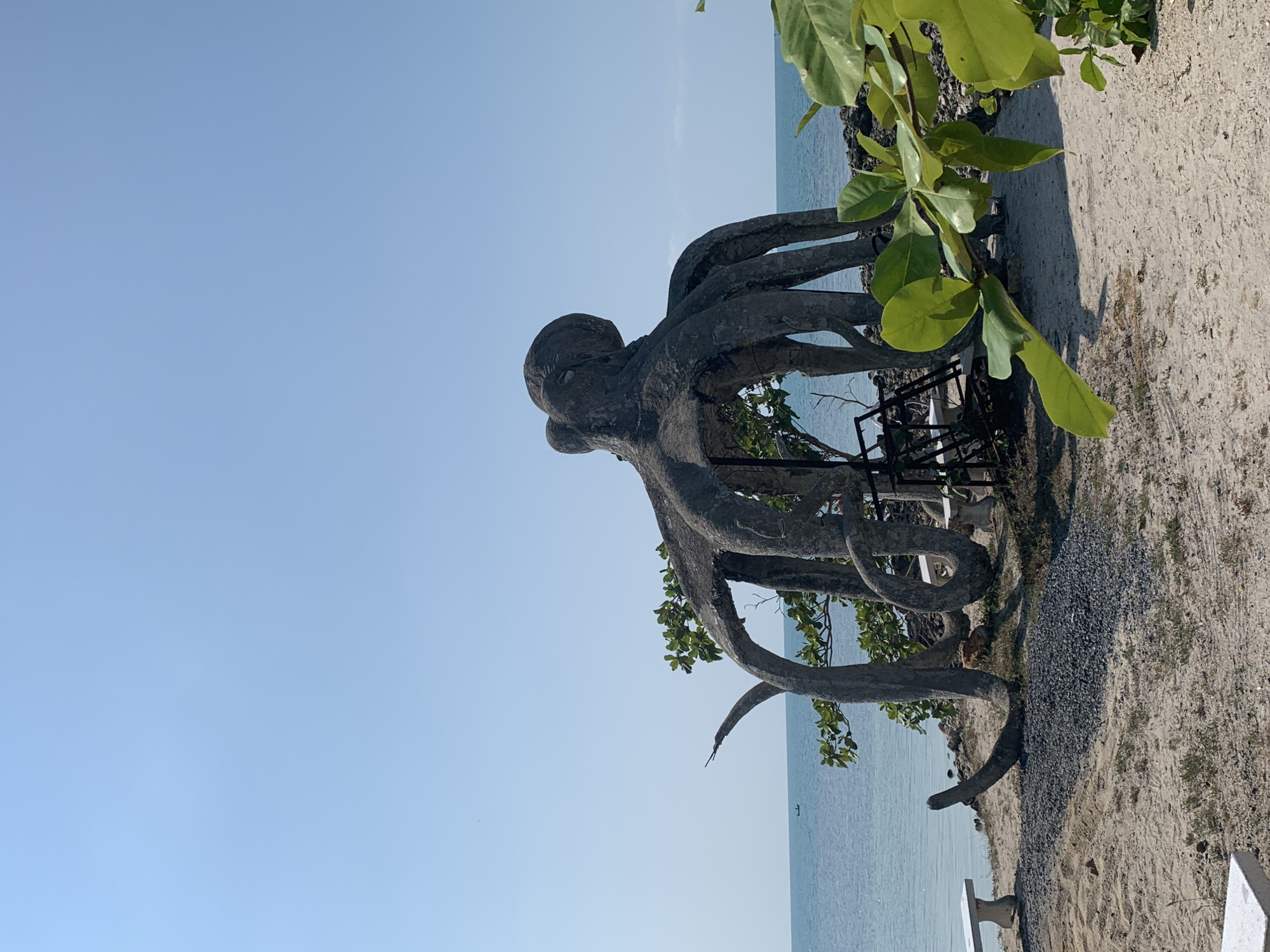 The image depicts an octopus statue located on the coast of Baan Tai, Thailand. The statue is made of gray concrete and has eight tentacles. It stands on a rocky outcrop covered in sand and gravel. The ocean is in the background, and the sky is clear and blue. Green foliage surrounds the statue, with a bench in the right-bottom corner of the image.