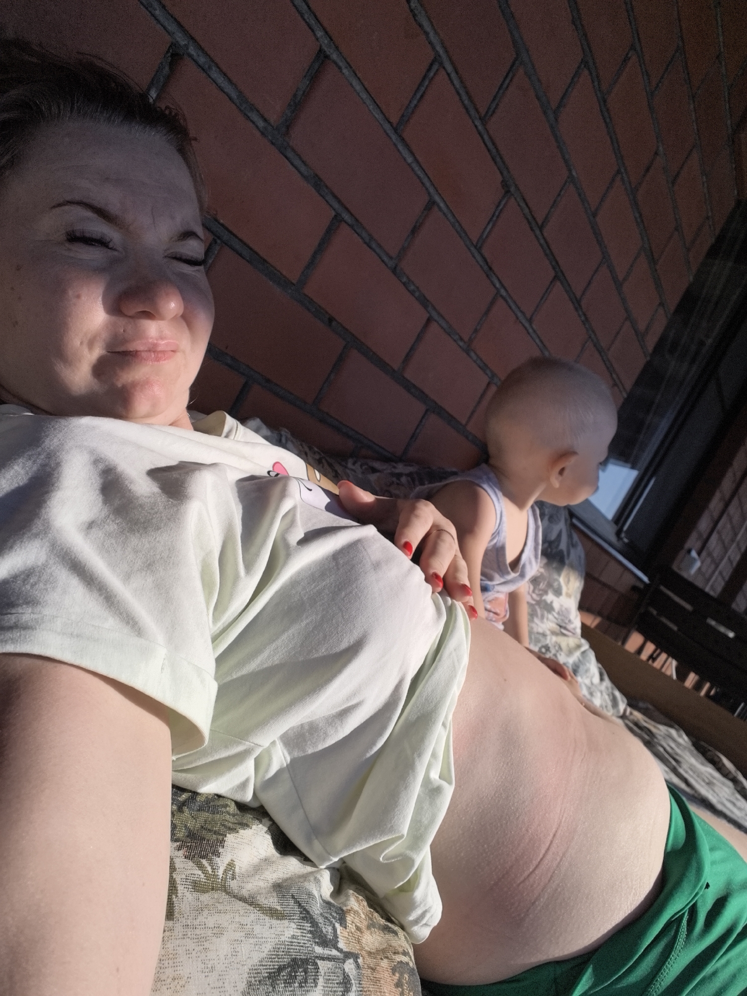 A medium shot from a low angle shows a woman lying down outdoors, with a baby sitting behind her. The woman is on the left of the frame, facing the camera with her eyes closed and a slight smile. She is wearing a light-colored t-shirt. Her abdomen is prominent and visible, with stretch marks apparent. She is wearing green shorts. The baby, on the right, is wearing a light blue tank top and is facing away from the camera, looking towards a window or balcony railing. The background consists of a brick wall and a patterned blanket or surface the subjects are lying on. The scene appears to be on a balcony or porch, possibly in Vityazevo, Russia, given the brick wall and the outdoor setting. The lighting suggests it is daytime, with sunlight casting shadows.