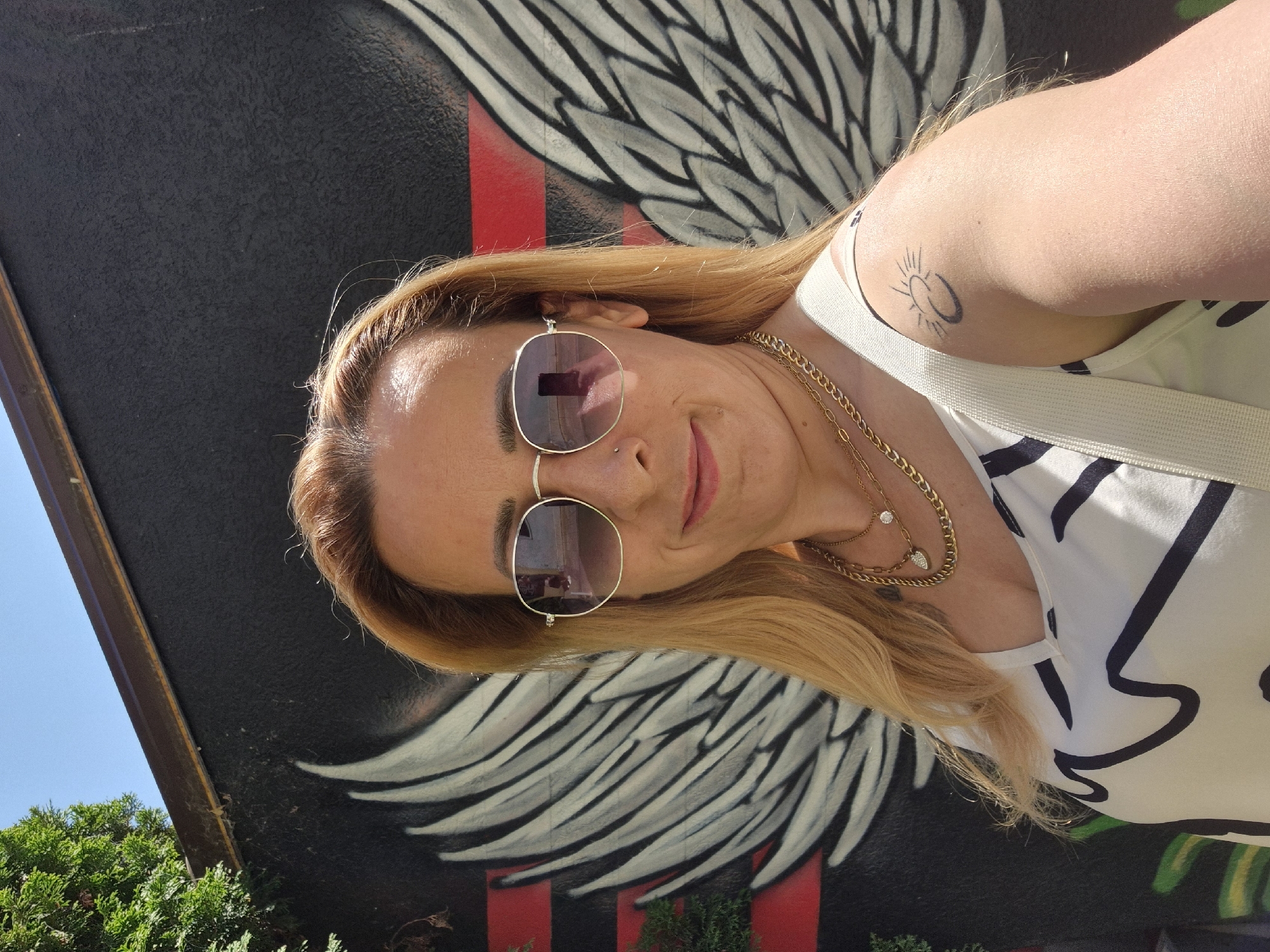 A woman with blonde hair and sunglasses stands in front of a mural featuring large, stylized white wings with red and black stripes. She is wearing a white top with black abstract designs and several gold necklaces. A faint tattoo of a sun with a crescent moon is visible on her right shoulder. The background also includes green foliage and a blue sky. The image appears to be a selfie, taken outdoors in a city.