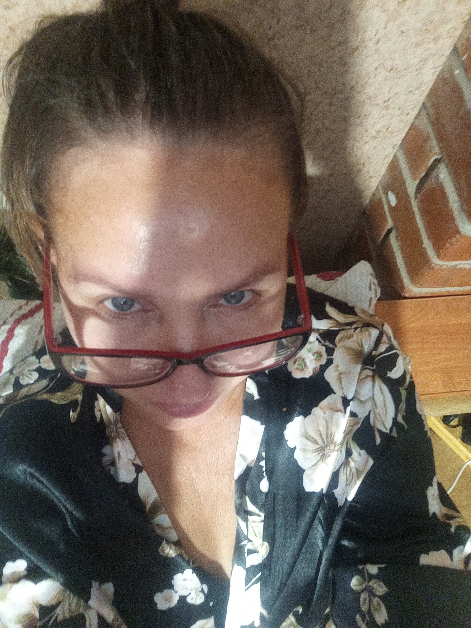 A woman is taking a selfie with a smartphone, as evidenced by the close-up perspective and the angle of her face. She is wearing glasses with red frames and a black robe with a floral pattern. The background shows a brick wall and a wooden surface, possibly part of a room in Novopokrovka, Russia. The lighting suggests an indoor environment.