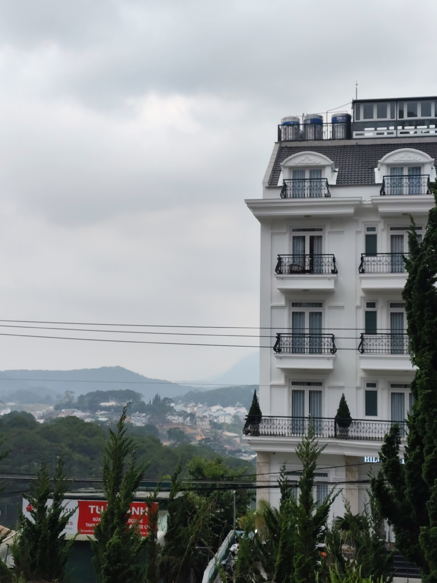 The image depicts a multistory white building with French-inspired architectural elements such as balconies with ornate railings and a mansard roof. The building is situated in the city of Cam Ly - Đà Lạt, Vietnam, as suggested by the style of architecture. The facade faces the viewer, revealing multiple floors with symmetrical window and balcony arrangements. The top of the building features what appears to be a rooftop structure and water tanks.
The background includes a landscape with hills and trees, and the sky is overcast. Utility wires are visible in the foreground. In front of the building, there are evergreen trees and part of a sign with Vietnamese script. The overall scene suggests a somewhat urban setting within a mountainous region. The lower right corner of the building has the name "HILL" visible, indicating the name of the hotel or building.