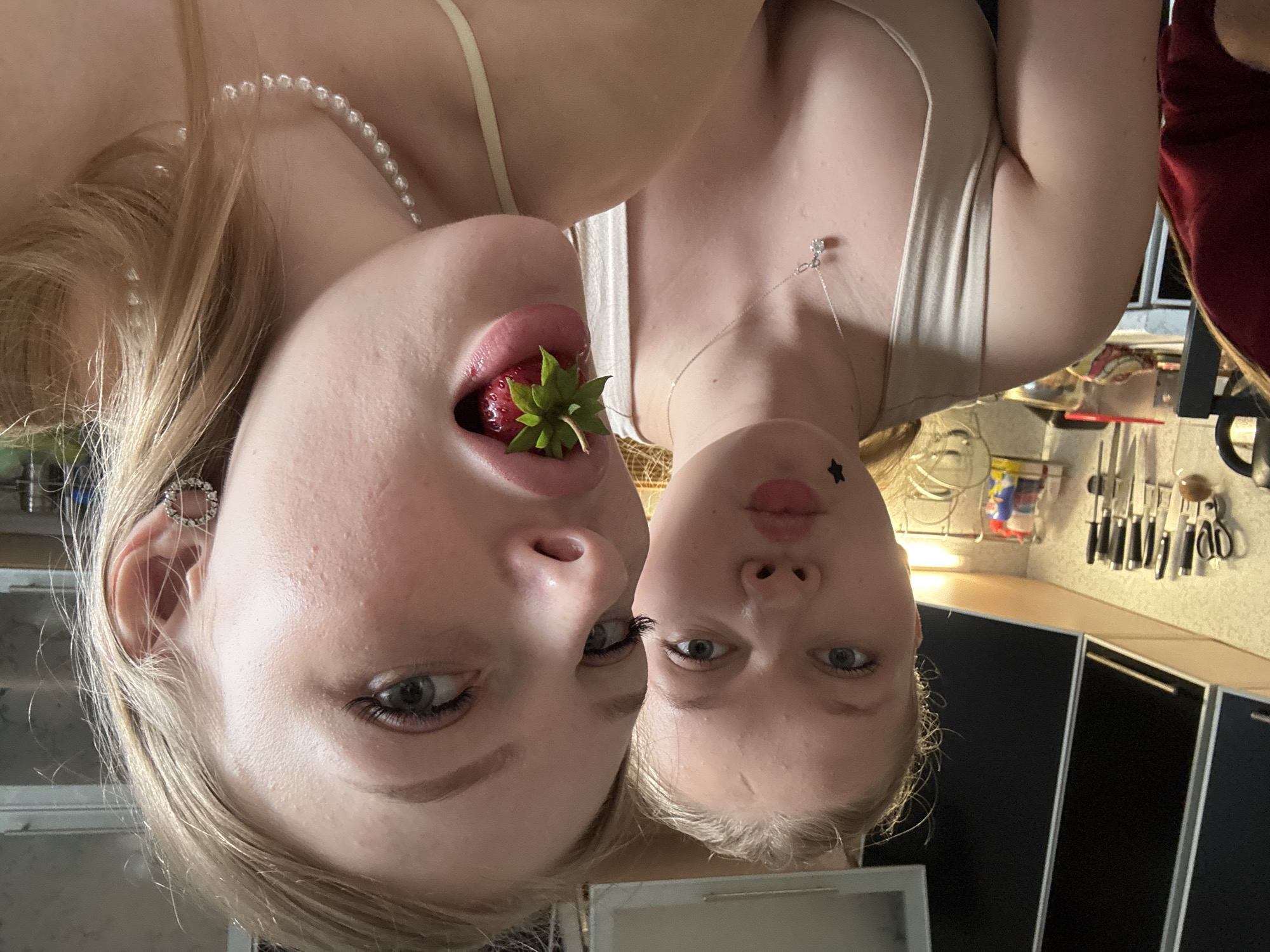 This is a photograph of two young Caucasian women in a kitchen, which appears to be indoors. The image is oriented vertically. One woman is in the foreground, leaning in towards the camera, with a strawberry in her mouth. She has blonde hair, light skin, and grey-blue eyes. She is wearing a pearl necklace and a sparkly earring in her left ear. The other woman is behind her, also leaning in. She has blonde hair, light skin, and grey-blue eyes. She has a star-shaped sticker on her left cheek and is wearing a silver necklace with a small pendant. Both women are wearing light-colored tank tops. The background shows kitchen cabinets and a knife rack on the wall, suggesting a domestic setting. The lighting appears to be artificial, typical of an indoor environment. The overall mood seems playful and intimate. There is no visible text in the image.