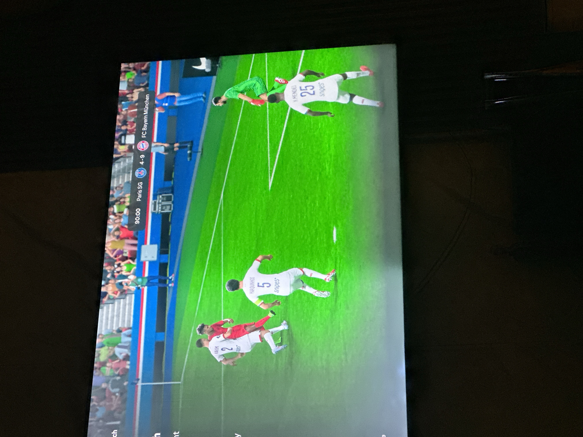 A football match is being played on a screen, displayed on what appears to be a television. The score indicates that "Paris SG" has 4 goals and "FC Bayern München" has 9 goals at the 90:00 minute mark. The players on the field are rendered in a video game style. The background shows a stadium with spectators. The scene is captured from an angle, suggesting it is being viewed in a room, possibly in a home or establishment in Gwarinpa, Nigeria, given the prompt.
