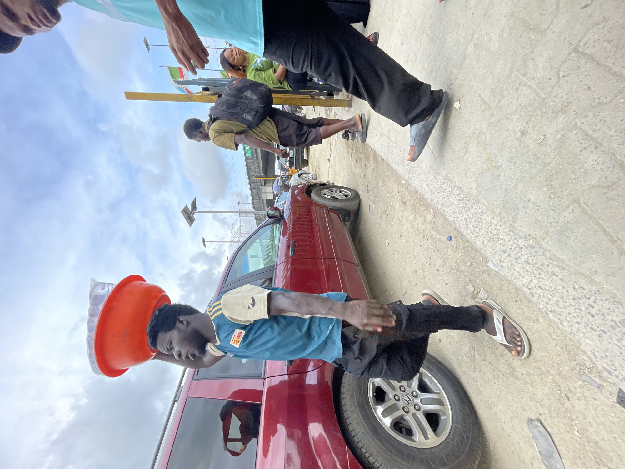 This rotated street scene, captured from a low, upward-looking perspective, depicts an active urban environment under an overcast sky. Several individuals are present, suggesting a bustling area, consistent with Lagos, Nigeria.
In the immediate foreground, the upper torso and head of a person wearing a light blue/teal shirt are visible on the left, with their arm extended into the frame. Next to them, the lower body of another person is seen, wearing black pants and grey sandals, standing on a paved surface.
Further back, a man in a yellow/olive shirt and dark shorts, carrying a black backpack, walks or stands. Behind him, a woman in a green top is visible, with her eyes closed or looking down.
A central figure is a man wearing a blue and beige short-sleeved shirt (possibly a jersey with "100 UNION" partially visible) and dark pants. He skillfully balances an orange bucket containing a plastic bag of what appears to be ice or water sachets on his head, indicative of a street vendor. He is positioned next to a red minivan or SUV, identified as a Honda CRV by its wheel hub.
The background features other vehicles, street poles with what seem to be solar panels, and flags with green, white, and red stripes. A green street sign partially reads "JUB," possibly an abbreviation for a street or landmark like "Jubilee." The ground is paved, suggesting a sidewalk or public walkway. The scene captures a moment of daily life in a vibrant city.
