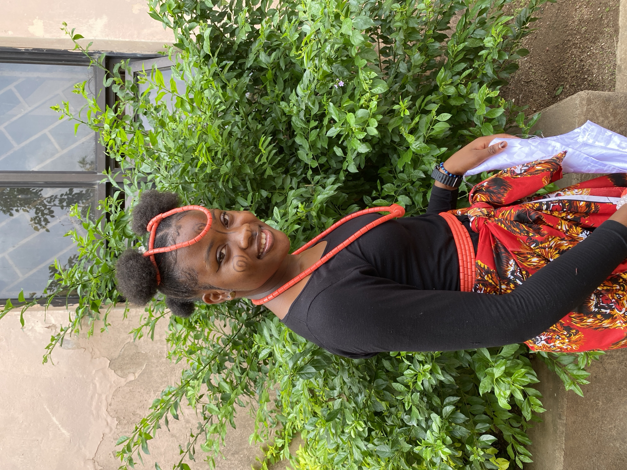 A young woman, likely in her late teens or early twenties, is posing for a photograph outdoors. She has dark skin and is wearing traditional attire, including a black long-sleeved shirt, a red and orange patterned wrapper, and coral beads around her neck and forehead. Her hair is styled in two puffy buns. Her expression is a smile, and she appears to be holding a white cloth in her right hand.
The background consists of lush green foliage. A window with a barred design is slightly visible in the upper left corner of the frame, and a beige wall is seen to the left of the foliage. The location is likely within the city of Gwong, Nigeria. The young woman has face paint.