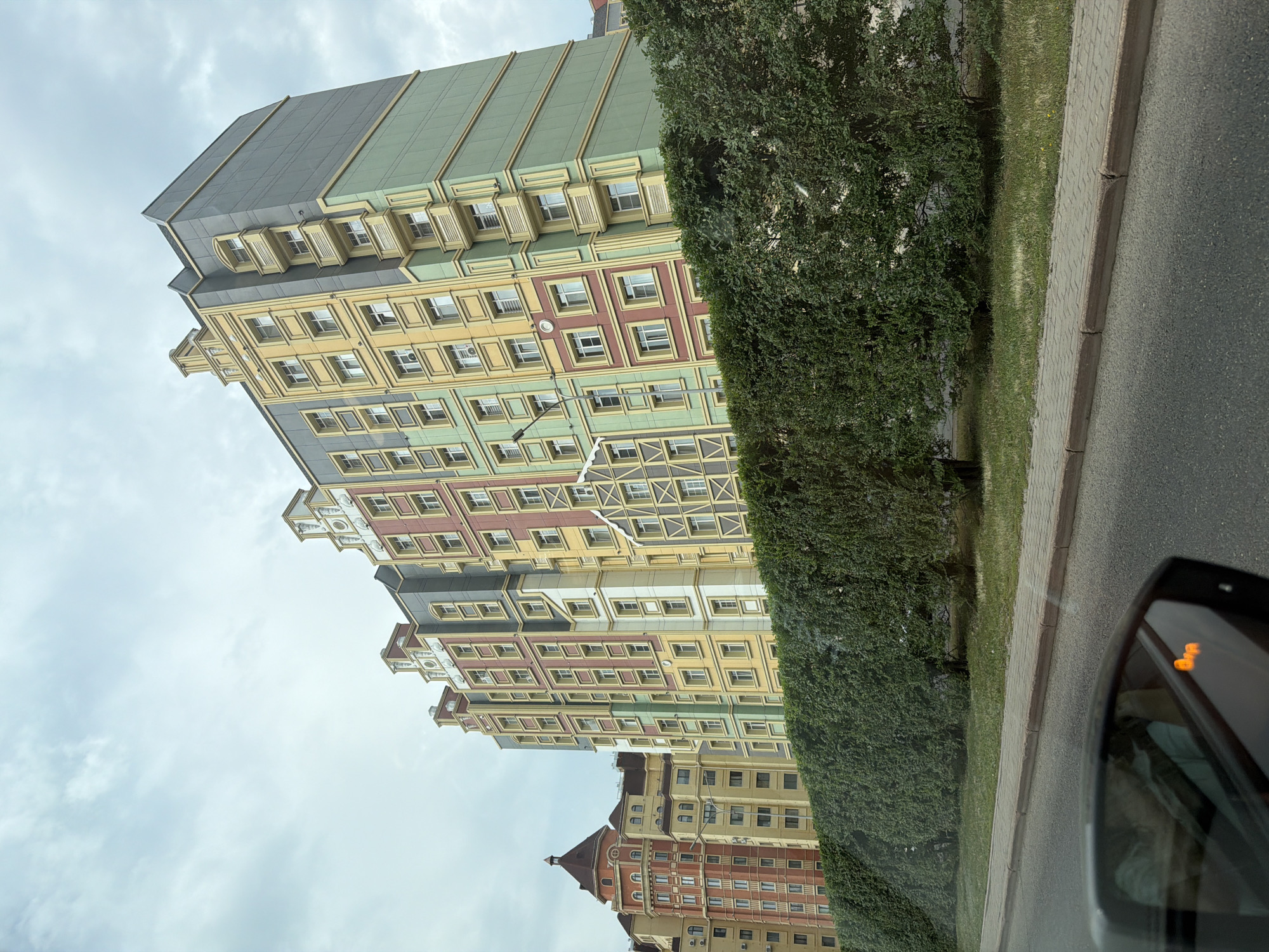 The image captures a street view in Astana, Kazakhstan, featuring a row of tall, colorful apartment buildings with distinctive stepped gables and varied facade colors, including yellow, pink, green, and gray. A dense, well-maintained green hedge runs along the front of the buildings, obscuring the lower portions. A paved sidewalk and an asphalt road are visible in the foreground, with the reflection of a person and a car's indicator lights visible in the car's side mirror on the left. The sky is overcast with clouds. The architecture of the buildings suggests a modern, perhaps themed, residential complex.