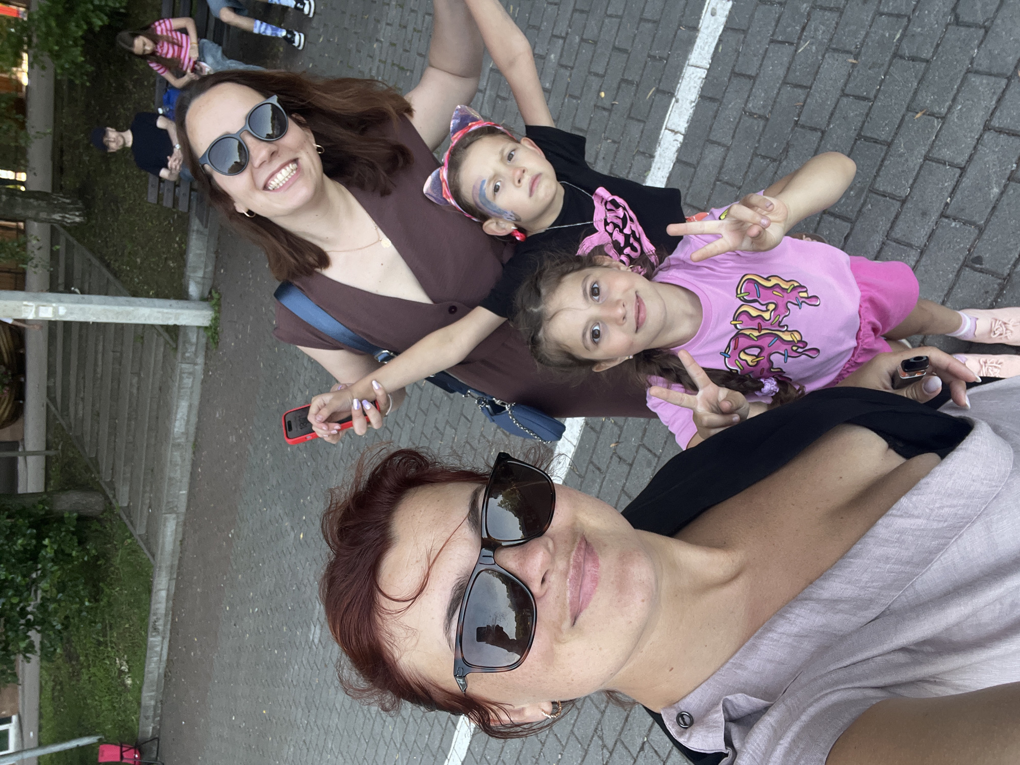 In Svetlogorsk, Russia, a group of four individuals, likely a family, are captured in a selfie taken outdoors on a paved surface, possibly a sidewalk or promenade, with trees and park benches visible in the background. The primary subject is a woman in the foreground wearing sunglasses and a light-colored top. To her right and slightly behind her is another woman, also wearing sunglasses and a brown dress, smiling at the camera and holding a red smartphone. Two young girls are positioned between them; one in a pink t-shirt with a donut graphic, the other in a black t-shirt with cat ears and face paint. The image appears to be taken during daylight hours, with natural light illuminating the scene. The overall atmosphere suggests a casual outing or gathering.
