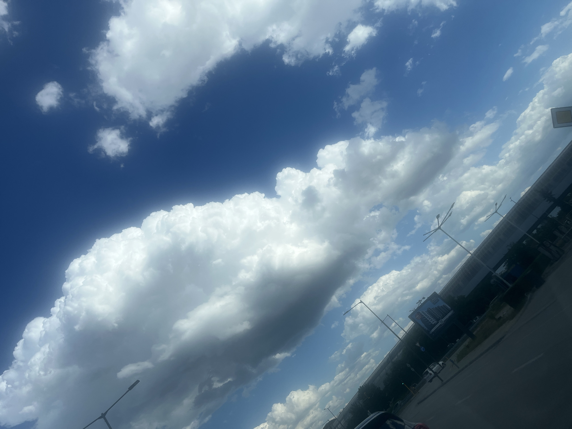 This is a tilted medium shot of a bright blue sky filled with large, fluffy white and gray cumulus clouds. In the lower left quadrant of the image, a road is visible, as are trees and a long, low-lying building or structure with a patterned roof. There are streetlights visible along the road and in front of the building. A large billboard is mounted on the side of the building. The overall scene suggests an outdoor environment, possibly a roadside area in a city or suburban setting.