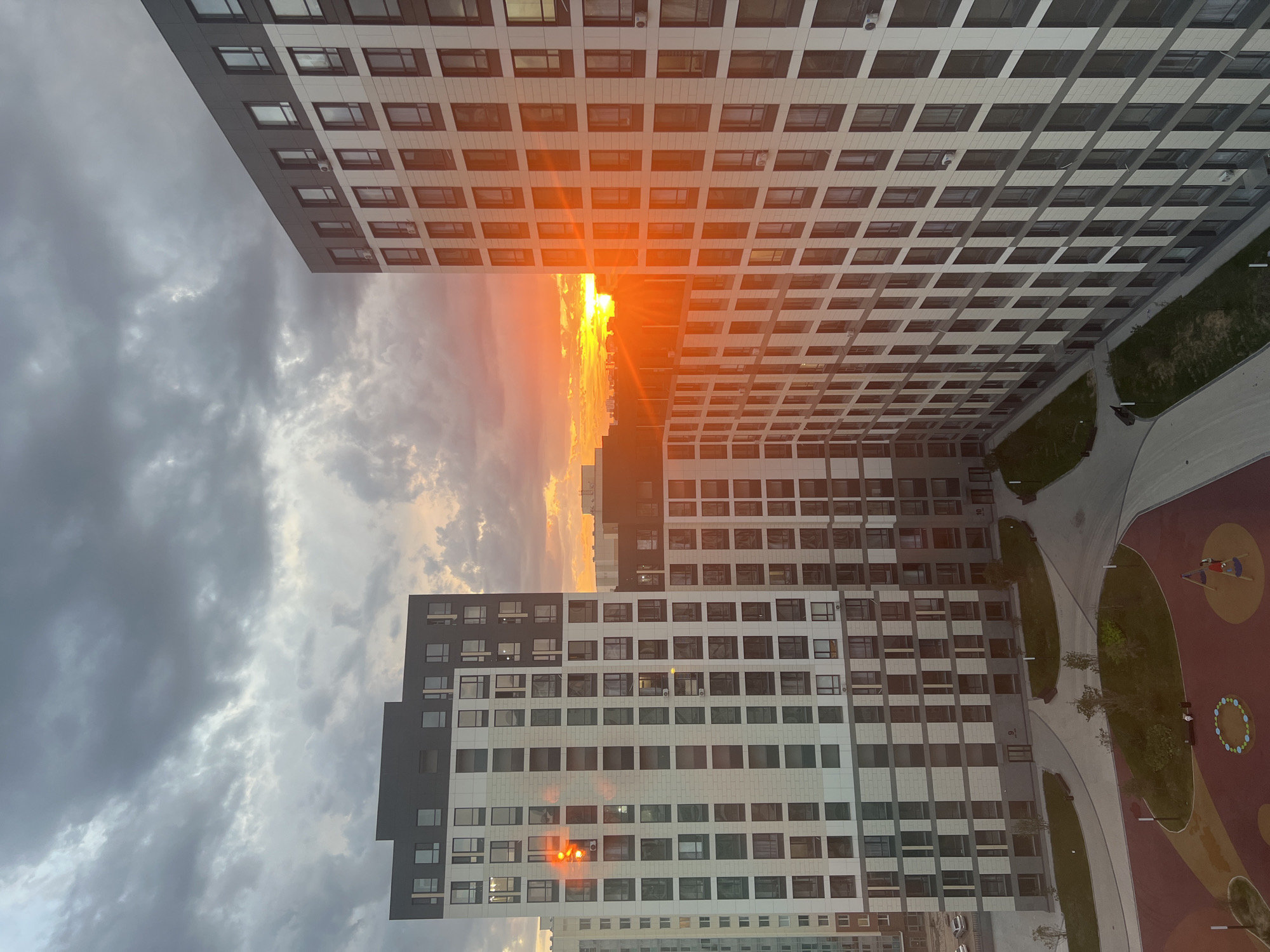 The image depicts a view of residential apartment buildings in Astana, Kazakhstan, during a sunset. The sky is filled with dramatic, dark clouds, with a vibrant orange and yellow sunset breaking through on the horizon, casting a strong orange glow on the buildings and creating lens flare effects. The architecture of the buildings is modern, characterized by clean lines, repetitive window patterns, and a combination of light-colored facades and darker accents. In the foreground, a courtyard area is visible with paved pathways, green spaces, and what appears to be a children's playground with colorful surfaces and play structures. There are no visible people in the scene. The overall impression is one of urban development under a striking natural phenomenon.