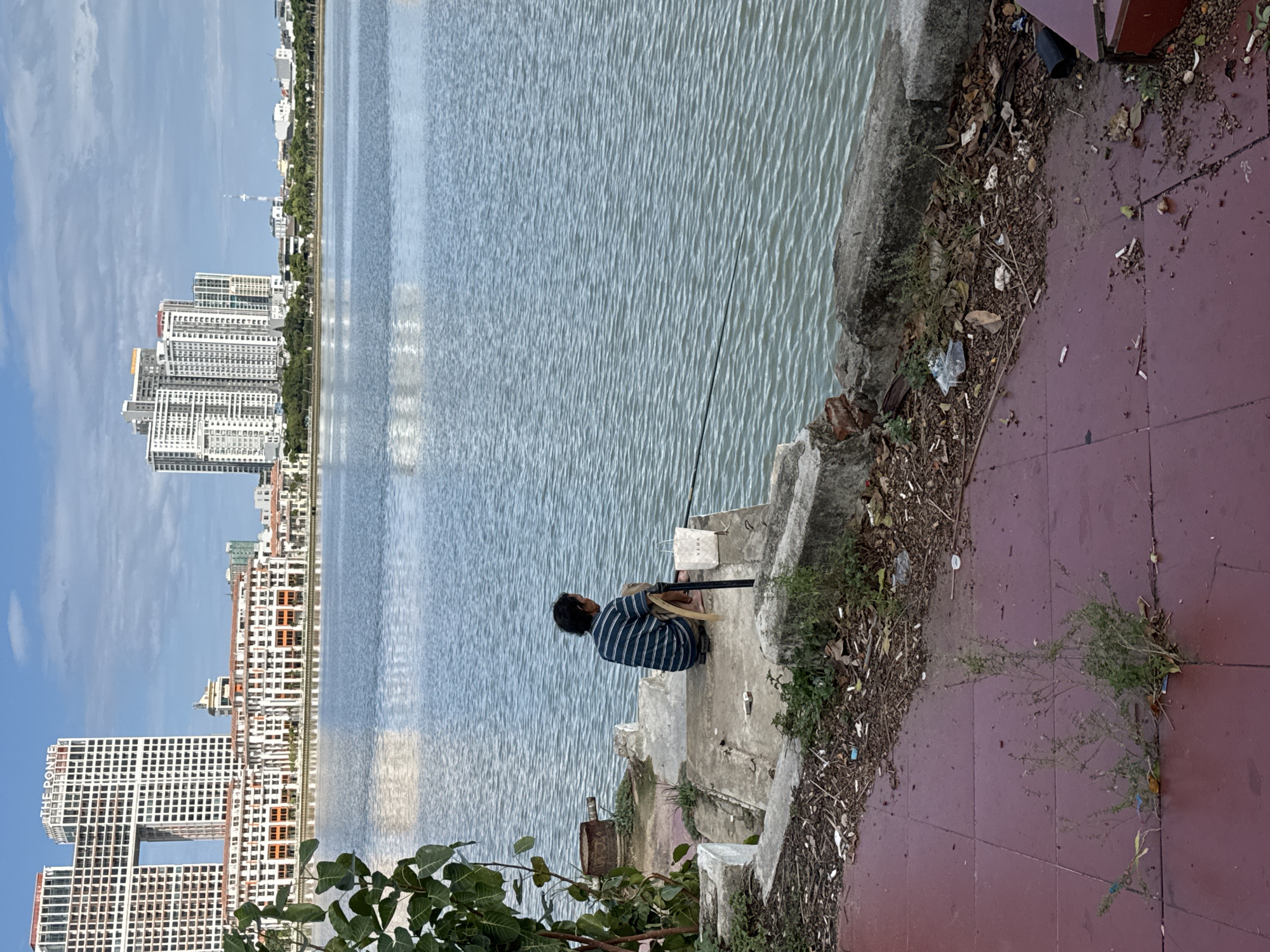 A lone individual, wearing a horizontally striped blue and white shirt and beige shorts, is seen from behind, sitting on a concrete ledge by a body of water, fishing with a rod. The person is positioned to the left of a white paper bag. The environment includes a waterfront area with a paved red walkway in the foreground, littered with debris including leaves, twigs, and plastic. Adjacent to the walkway is a concrete embankment and then the calm, rippled water. In the background, across the water, a cityscape unfolds with modern, multi-story buildings of varying heights and architectural styles. Prominent structures include a white building with a large rectangular opening near its top and another cluster of tall, white residential or office towers. The sky above is partly cloudy. The scene appears to be located at a lakeside or riverfront area in the city of Đà Nẵng, Vietnam, given the architectural style of the buildings.