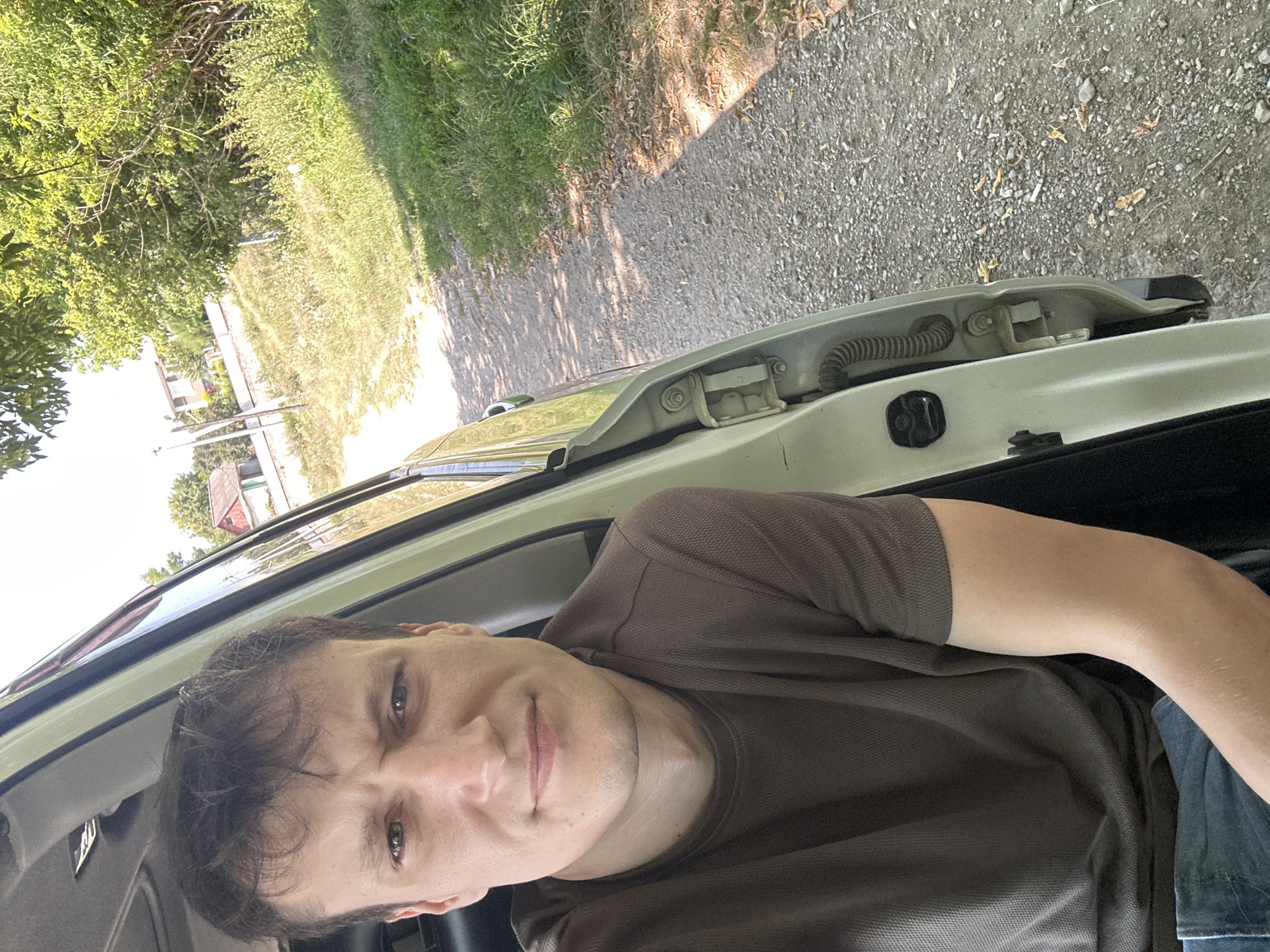 A close-up, high-angle, selfie-style shot shows a man in his late 20s to early 30s sitting inside a light-colored vehicle, with the driver's side door open. He has light brown hair, fair skin, and green eyes, and he is wearing a brown t-shirt and blue jeans. His expression is neutral. The background shows a dusty, unpaved road with a grassy embankment on the right, and some trees and buildings in the distance. The scene is consistent with a rural area, possibly on the outskirts of Makiv, Ukraine.