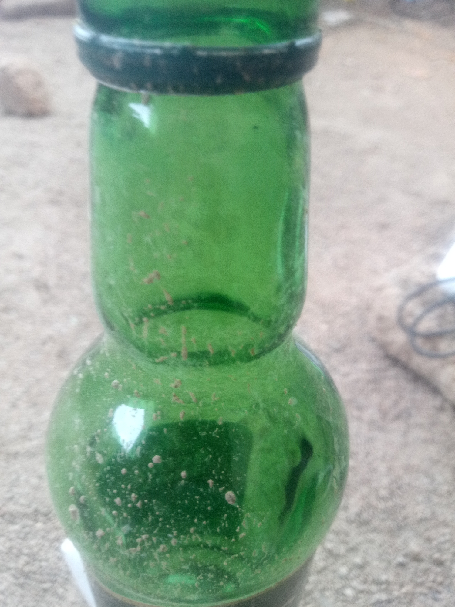 A close-up, eye-level shot shows a dirty green glass bottle. The bottle has a bulbous bottom section, tapering to a narrower middle section, and then widening again towards the top, which is obscured. The glass is covered in small, reddish-brown specks and some larger blurry specks, suggesting it is either old or has residue on it. The background is out of focus and appears to be a sandy or gritty surface, possibly ground or a rough outdoor texture. A blurred object, possibly a coiled wire or cable, is visible in the upper right corner. There is also a blurry rock-like object in the upper left corner. The lighting is natural and diffuse, casting soft shadows.