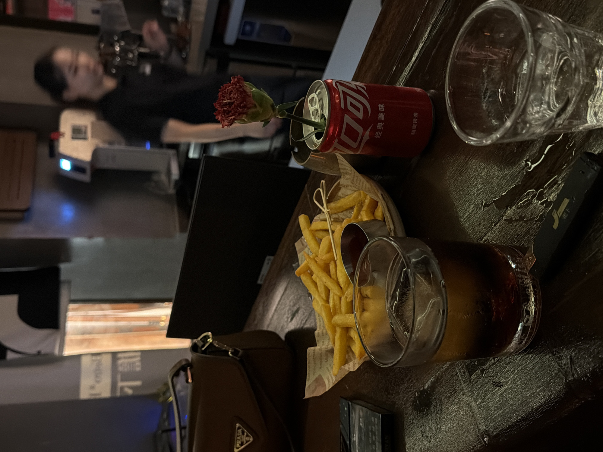 The image depicts a dimly lit indoor scene, likely a bar or restaurant in Longgang District, China. In the foreground, a wooden table is laden with various items. A glass of dark liquid with ice sits on the left, next to a pack of cigarettes. In the center, a basket of french fries is accompanied by a small metal container of ketchup. A red Coca-Cola can, used as a vase for a single red carnation, stands to the right of the fries. Further to the right, another empty glass is visible. In the background, a woman, presumably an employee, is blurred in motion behind a counter, with a coffee machine and other bar equipment visible. A brown handbag with a Prada logo rests on the left side of the table, partially obscuring a wall with illuminated signage. The overall ambiance is casual and suggests a relaxed evening setting.