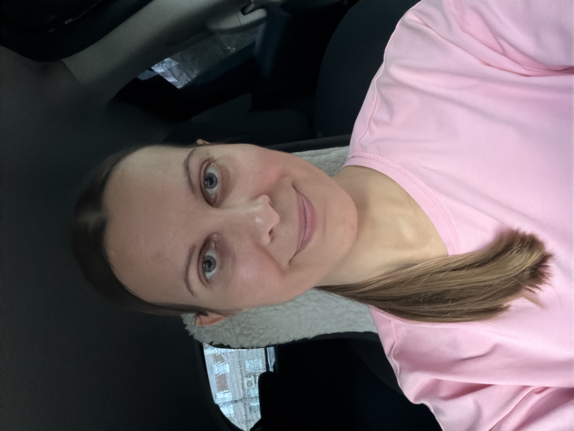 This is a selfie taken by a woman in a car. She has light skin, blue eyes, and brown hair pulled back into a ponytail. She is wearing a pink t-shirt. The background is dark, suggesting the interior of a vehicle, with a hint of a fuzzy material, possibly a seat cover or lining, and what appears to be a window with buildings visible outside, possibly in Izhevsk, Russia. The lighting is dim, but her face is well-lit, and she has a slight smile, conveying a relaxed or pleased emotion. The image is oriented sideways.