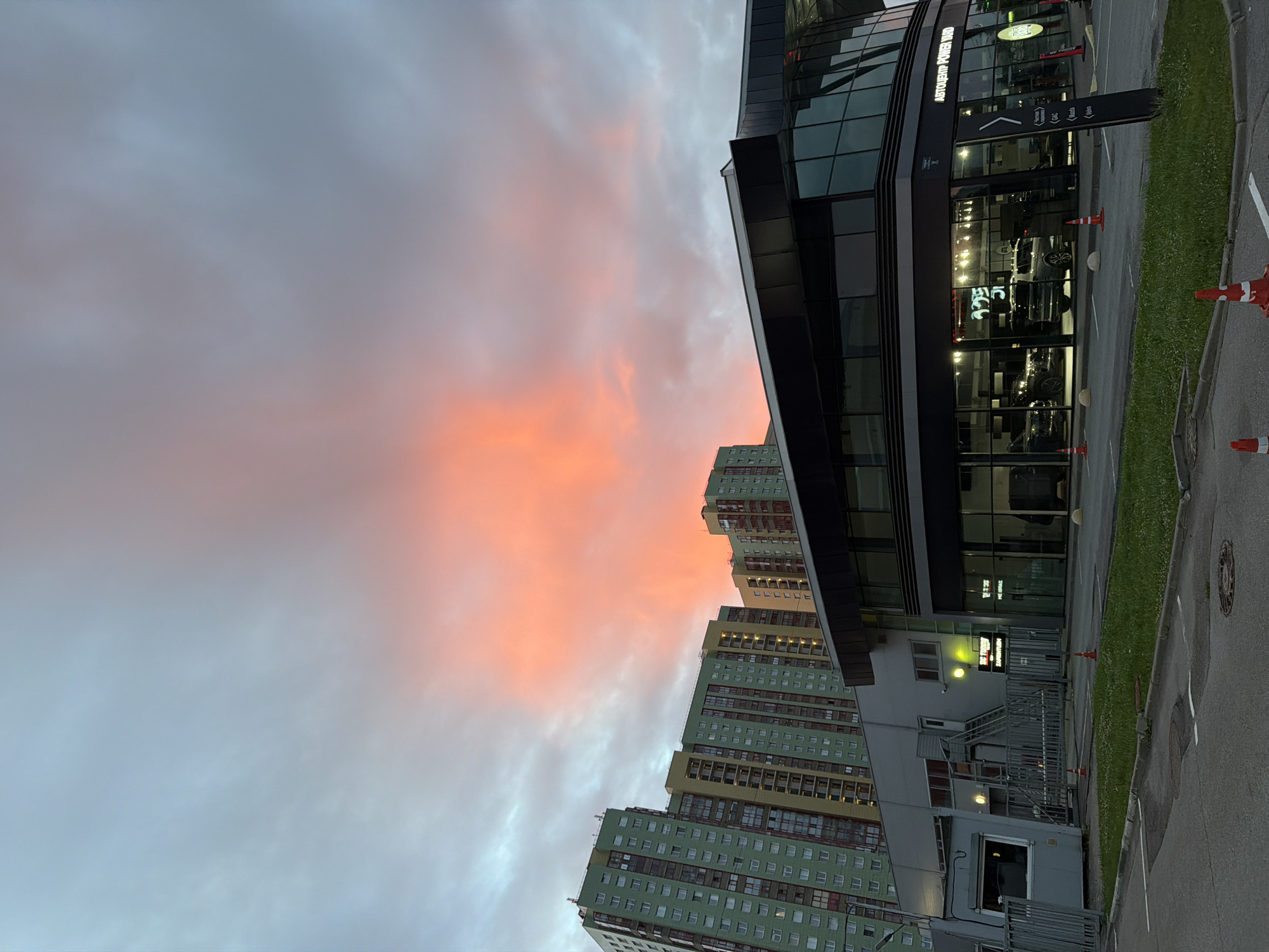 A wide shot captures a modern, multi-story building with a dark, angled facade featuring large glass windows, presumably a car dealership, at the forefront. Inside, several cars are visible. To the left, a cluster of tall, colorful residential buildings with green and yellow facades dominate the skyline. The sky above is dramatic, with a large portion illuminated by a vivid orange and pink sunset, contrasting with the cooler blue and gray tones of the clouds. The scene is set in a city, as indicated by the urban architecture. The ground in front of the dealership appears to be an asphalt lot with patches of grass, some traffic cones, and a manhole cover. The lighting suggests it is either dawn or dusk.