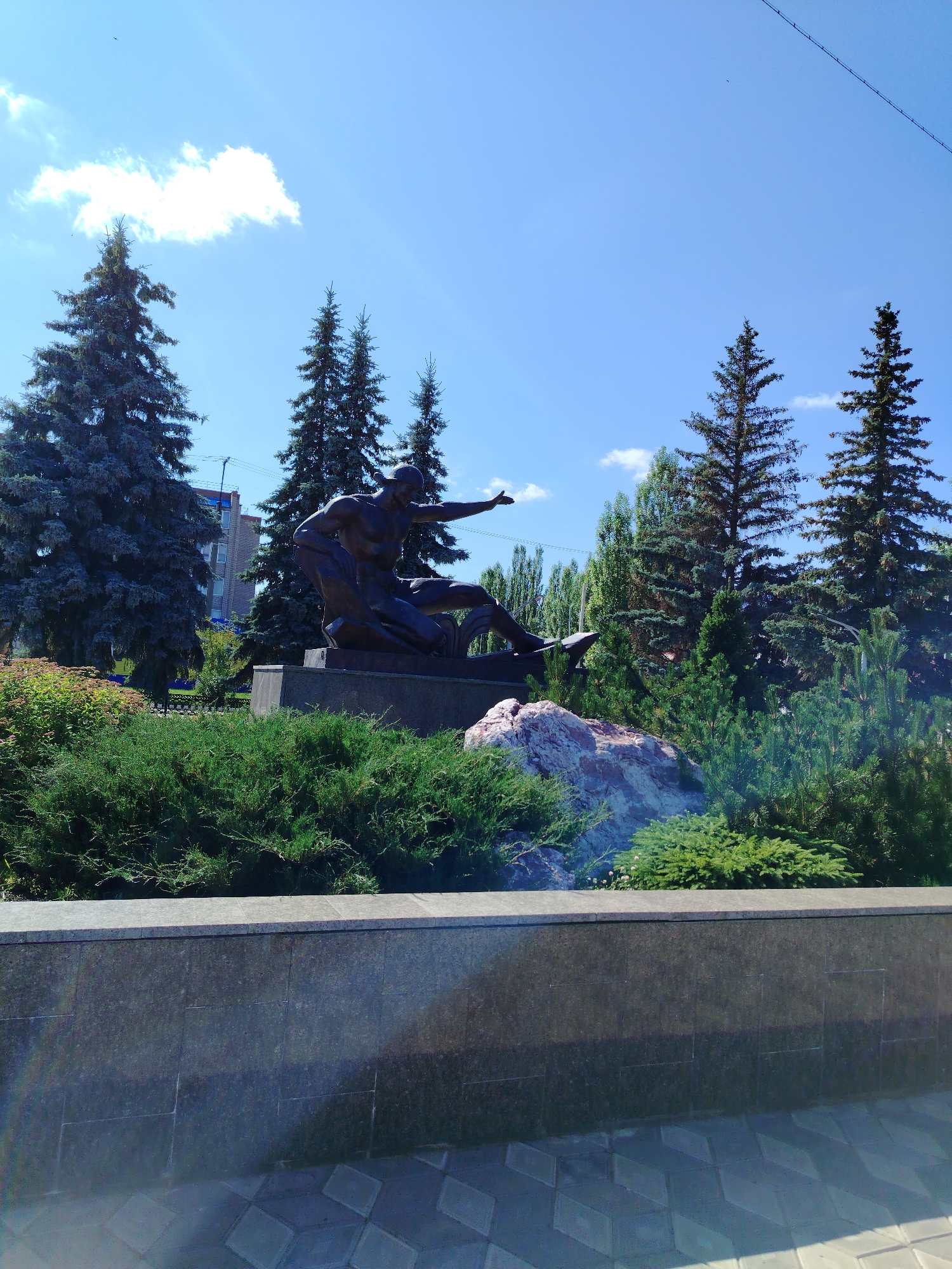 A bronze statue of two men is displayed outdoors on a sunny day. The statue is situated in front of a backdrop of tall evergreen trees and a clear blue sky with scattered clouds. In the foreground, a low stone wall and lush green bushes lead the eye towards the monument. A large, light-colored rock sits to the right of the statue, nestled amongst the foliage. A building with multiple windows is visible behind the trees on the left side of the frame. The ground in front of the wall is paved with geometric tiles. This scene is located in the city of Yanaul, Russia.