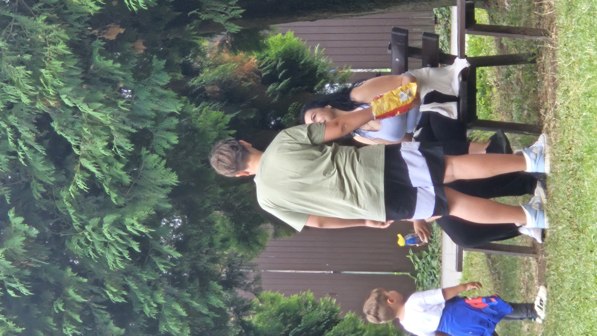 The image depicts an outdoor scene, likely a park or backyard, with lush green foliage dominating the background. In the foreground, a young boy in a white t-shirt and blue jacket stands on the left, facing away from the camera. To his right, a young man in a light green t-shirt and black and white shorts is also facing away from the camera. He is holding a bottle. Seated on a wooden bench in the center is a woman wearing a light blue top and white pants. She is holding a bag of chips. The woman appears to be interacting with the young man, possibly looking at him. The ground is covered in grass. The overall impression is a casual outdoor gathering. The specific location in Prešov, Slovakia is not determinable from the visual information provided.