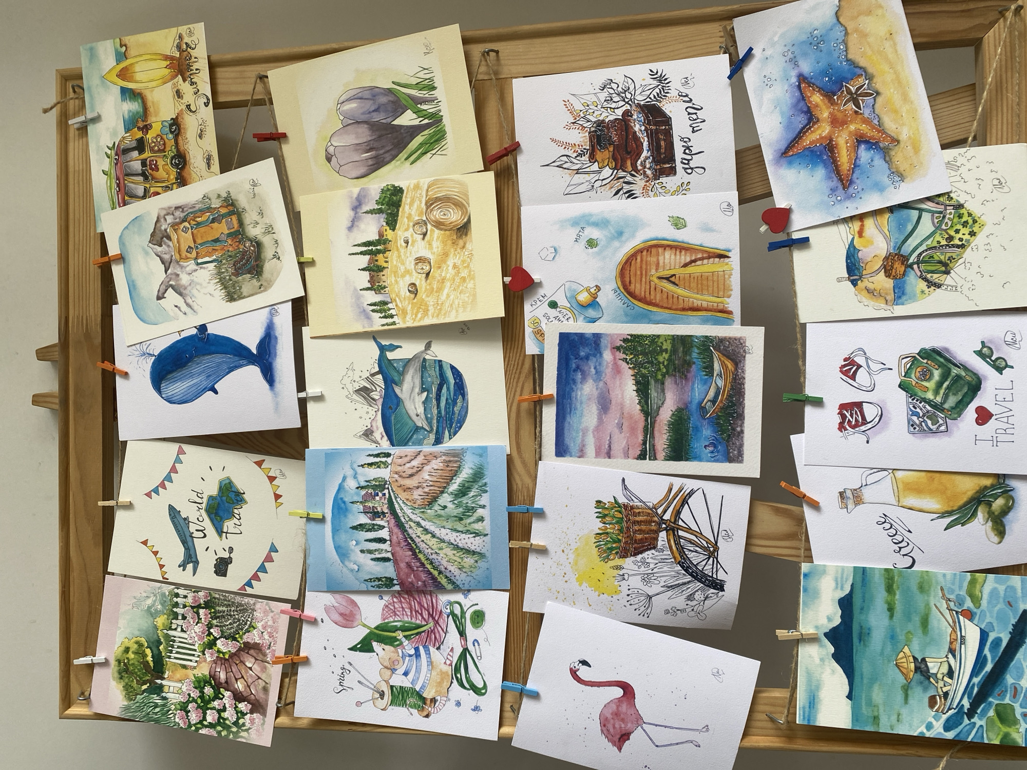 A wooden frame displays a collection of approximately twenty postcards attached with clothespins. The postcards feature various watercolor illustrations, including travel-themed images (camper vans, suitcases, maps), nature scenes (flowers, trees, whales, starfish, flamingos), food items (olives, olive oil), and other whimsical subjects (a cat sewing, a boat, abstract landscapes). Text and lettering in various languages are visible on some of the postcards. The overall aesthetic is artistic and handmade. The location in Yekaterinburg, Russia, is not directly evident from the visual information in the image.
