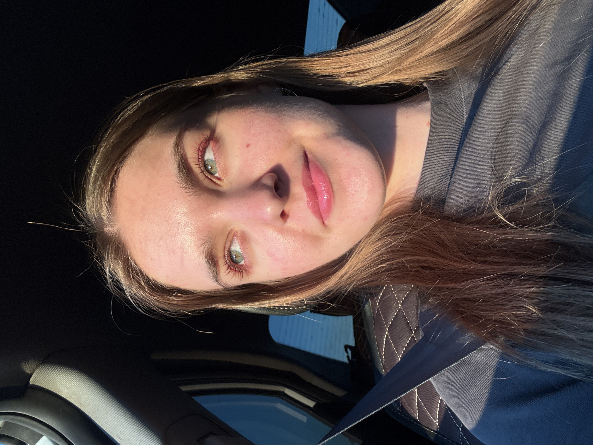 A young woman with light brown hair and green eyes is sitting in the passenger seat of a car, looking to her right. She is wearing a dark gray t-shirt and has pink lipstick on. The sun is shining through the window, casting a shadow across her face and the interior of the car. The car's interior is dark, with a dark gray seat visible behind her and a seatbelt across her chest. The location is implied to be Ustinovo, Russia, although no specific landmarks are visible.