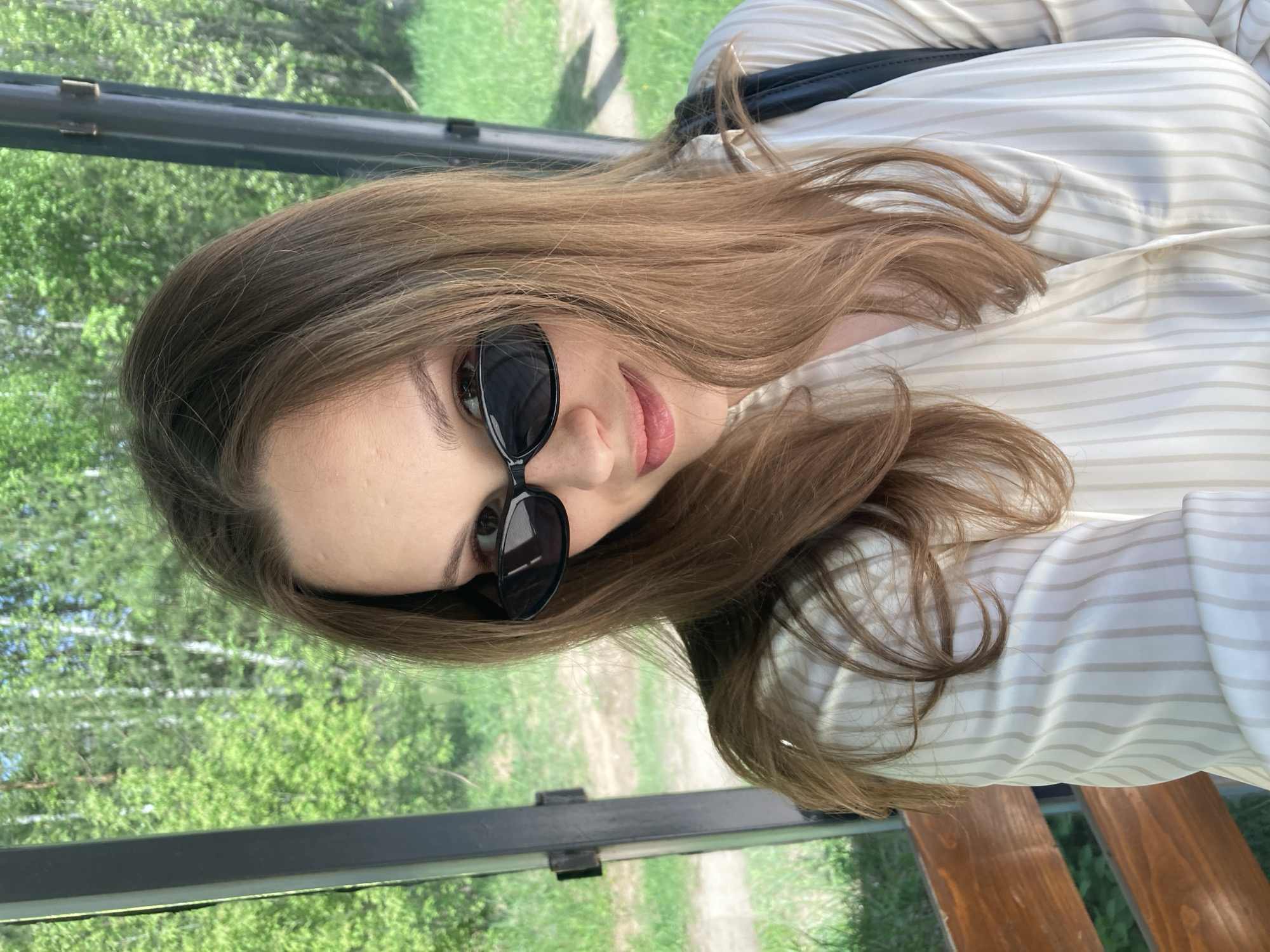A woman wearing sunglasses is the primary subject of this medium-shot photograph. She has long, light brown hair and is wearing a white, striped button-down shirt. Behind her, through what appears to be a glass or open-air structure, a green, wooded area is visible. The woman is indoors, possibly sitting on a bench or seat, as indicated by the wooden slats visible at the bottom right of the frame. The location is suggested to be Chaikovsky, Russia, although no specific landmarks confirming this are visible in the image.