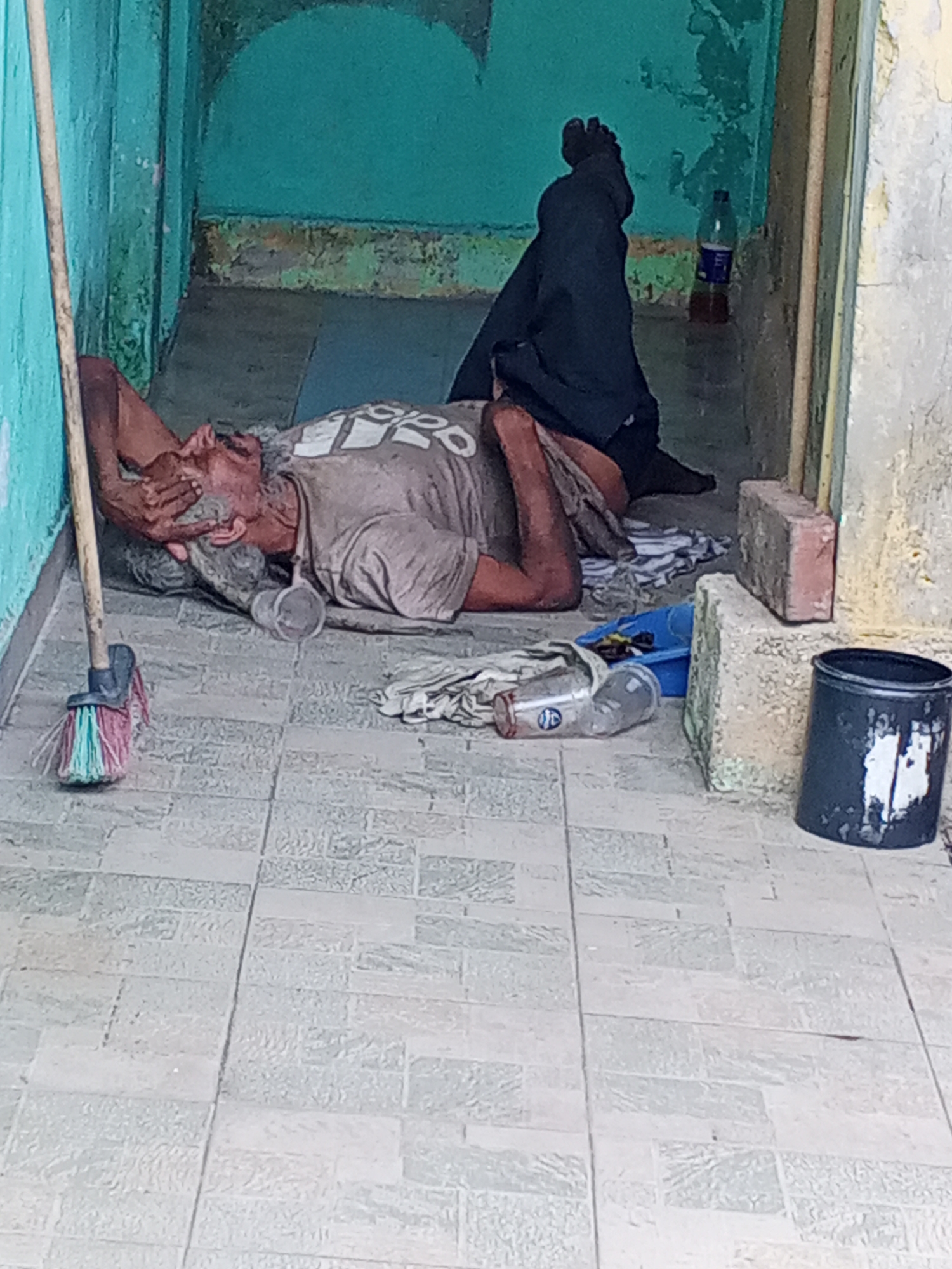 A man lies prone on a tiled floor inside a building in Tinaquillo, Venezuela. He is lying on his side with his face obscured by his hand. The man is wearing a t-shirt and dark pants, with his bare feet visible. A plastic cup, plastic bottle, bucket, and a broom are nearby. The walls of the building are painted in shades of blue and are weathered. A water bottle sits in the background. The setting is unsanitary and possibly abandoned.