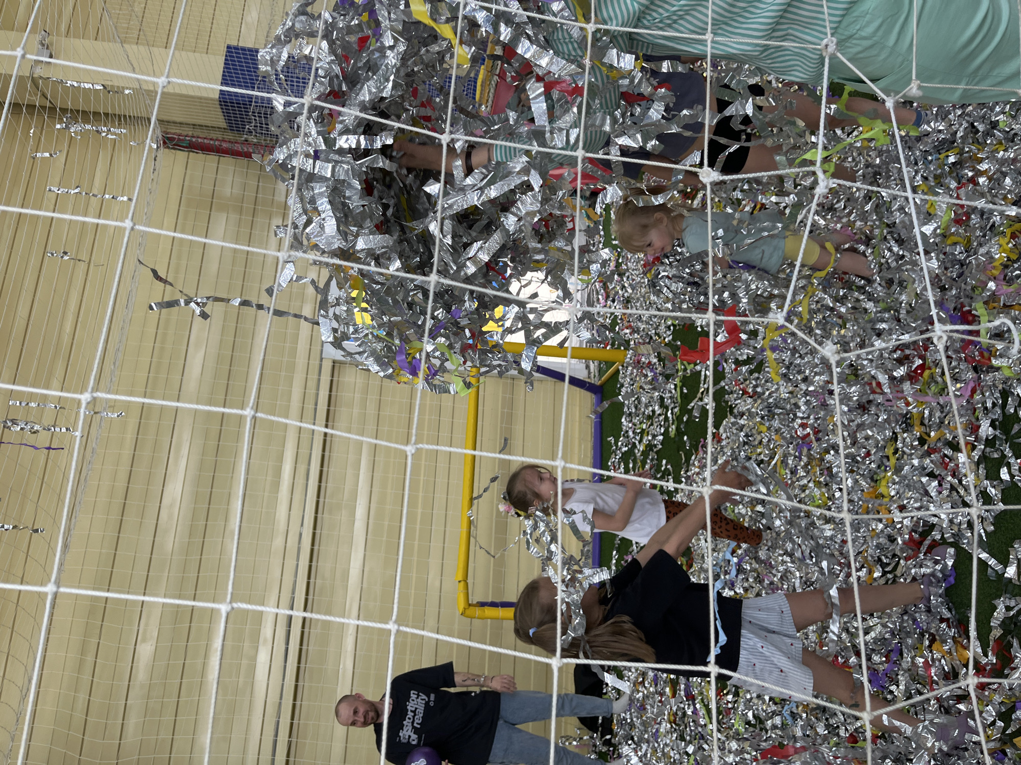 A partially obscured view through a white net reveals a scene inside what appears to be an indoor playground or recreational area. The scene is set in the city of Chaikovsky, Russia. A layer of silver and colorful confetti covers the ground, suggesting a celebration or event.
Several people are present, including two young children. One child, seen towards the top of the image, is lying on the confetti. Another child is wearing a white top. Further down in the image is a girl with long blonde hair, wearing a black t-shirt and shorts.
In the lower left corner of the image, a man is visible, wearing a black t-shirt.
A yellow frame structure is visible. The overall scene suggests a playful atmosphere.
