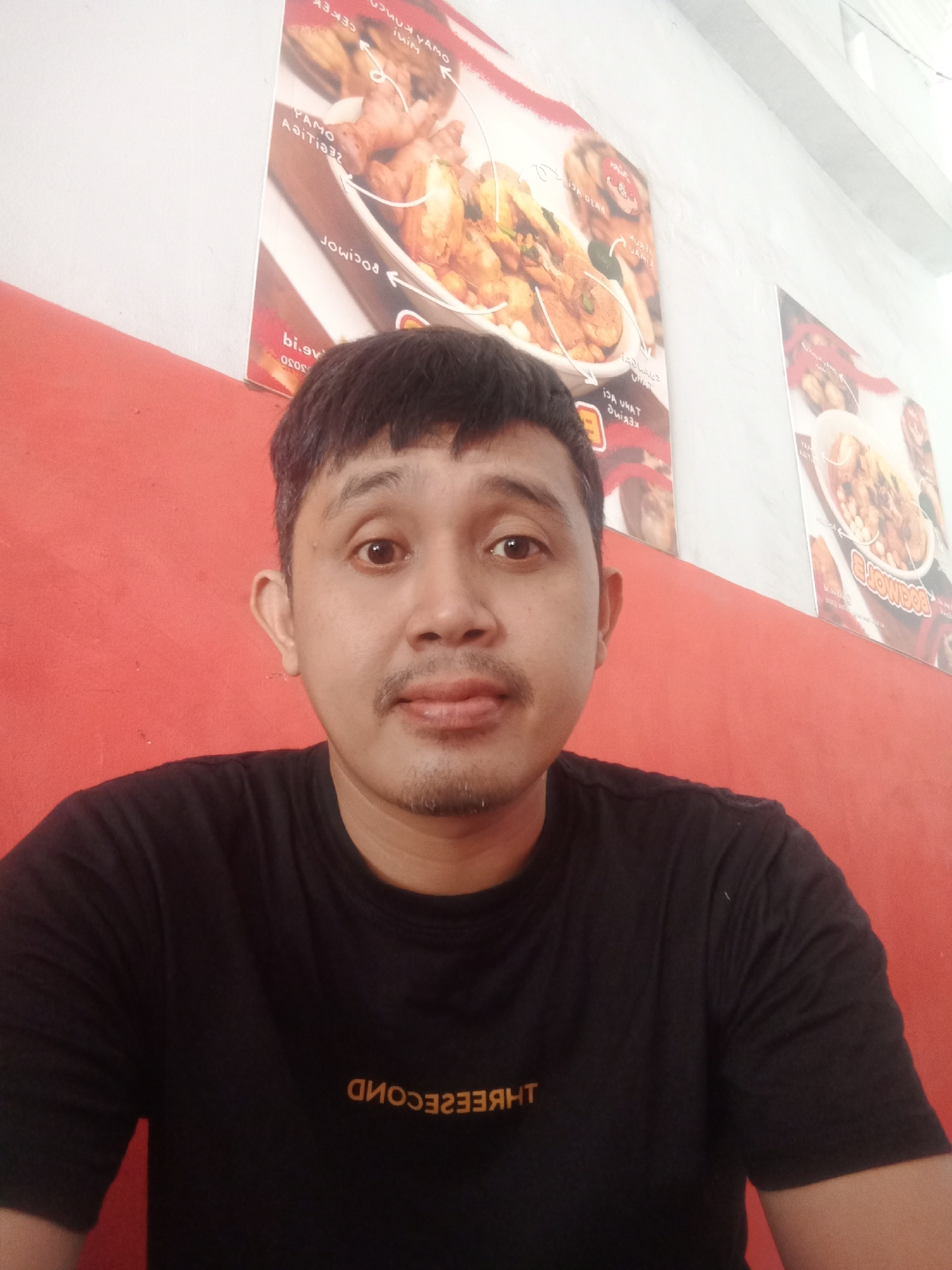 A middle-aged Asian man is seated indoors, likely in a restaurant, as evidenced by the food posters behind him. He is wearing a black t-shirt with the word "THREE SECOND" printed on it. The background consists of a red wall and a white wall. Two food posters are visible on the white wall. The man is looking directly at the camera with a neutral expression. This photo was taken in Kaliwungu, Indonesia.