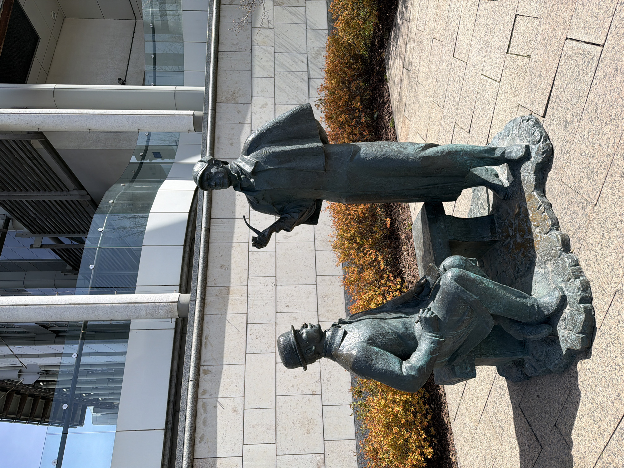 This image captures a bronze sculptural group depicting two male figures, widely recognized as Sherlock Holmes and Dr. Watson, set outdoors in an urban environment in Moscow, Russia.
The standing figure, identifiable as Sherlock Holmes, is shown wearing a deerstalker hat and a long coat. He holds a pipe in one hand, appearing to be in a moment of contemplation or observation. Beside him, Dr. Watson is seated on a sculpted bench-like element, dressed in a bowler hat and suit, with what appears to be a notebook or journal on his lap, suggesting he is recording information. Both statues are rendered in a classic, detailed style with a greenish patina characteristic of aged bronze.
The sculptures are situated on a light-colored paved area, resembling a plaza or walkway with geometric tiles. Behind the statues, a low hedge or shrubbery with autumn-toned foliage (yellows and oranges) provides a natural backdrop. In the background, a modern building with extensive glass windows and light-colored paneling reflects the bright sky and surrounding structures, creating a contemporary urban setting.
The strong, distinct shadows cast by the statues on the pavement indicate a clear, sunny day, likely during midday or early afternoon. The overall scene conveys a sense of intellectual engagement and quiet observation, fitting the characters. No visible text is present on the statues or in the background.