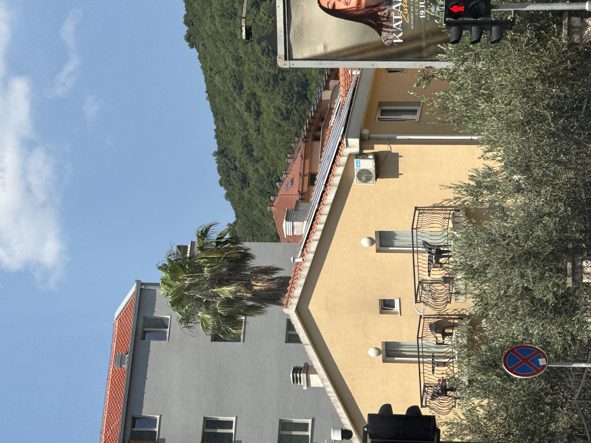 In Budva, Montenegro, a bright, sunny day is captured with a clear blue sky and scattered clouds. The foreground is dominated by lush green olive trees, partially obscuring a street scene. A traffic light displays a red pedestrian signal, and a circular sign with a red border and a blue 'X' indicates a parking restriction.
Behind the trees, a yellow building with balconies and solar panels is visible. Two balconies feature wrought-iron railings and are furnished with chairs and tables. An air conditioning unit is mounted on the side of the building. Further back, a taller, grey building with a red-tiled roof stands, featuring several windows and a prominent chimney. A tall palm tree with a thick trunk and green fronds is situated between the two buildings.
In the background, a densely forested green hillside rises towards the sky. To the right of the scene, a large billboard featuring a woman's face is partially visible, along with some text that appears to be event information. A street lamp arm extends from the upper right corner of the image.