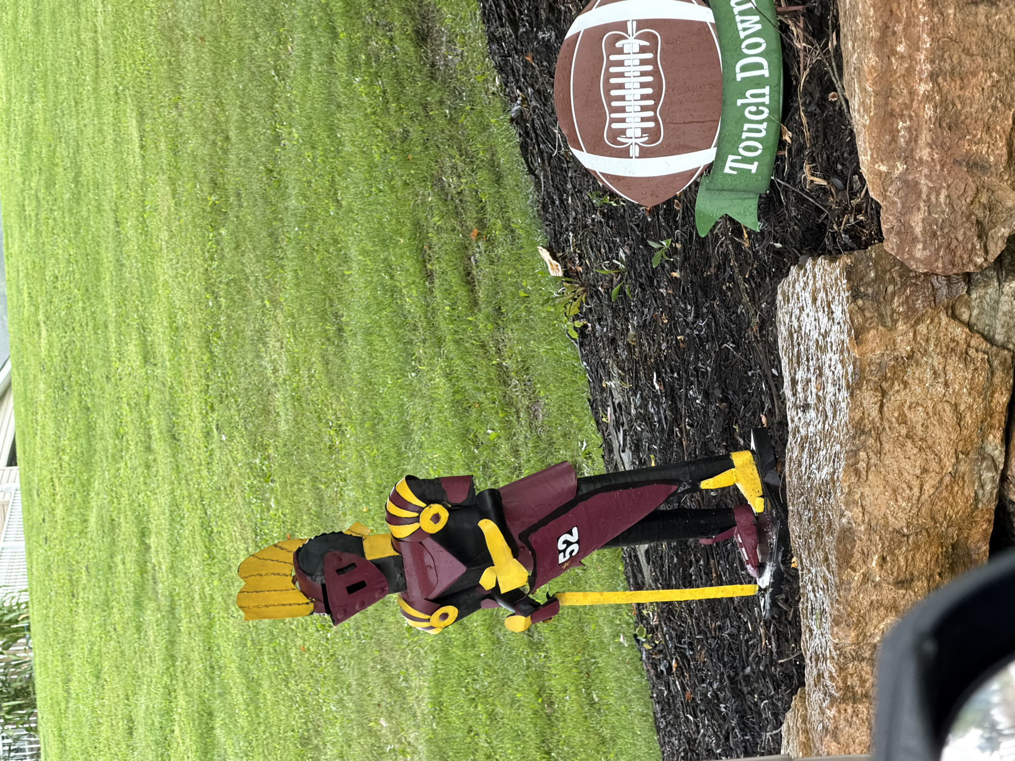 In West Pike Run Township, USA, an outdoor scene features a decorative wooden knight figure standing on mulch. The knight is painted in burgundy, black, and yellow, with the number "52" visible on its armor. It holds a yellow pole and wears a helmet. To the right of the knight is a wooden cutout of a football with the words "Touch Down" on a green banner below it. The figures are situated in front of a grassy slope and behind a large rock formation.