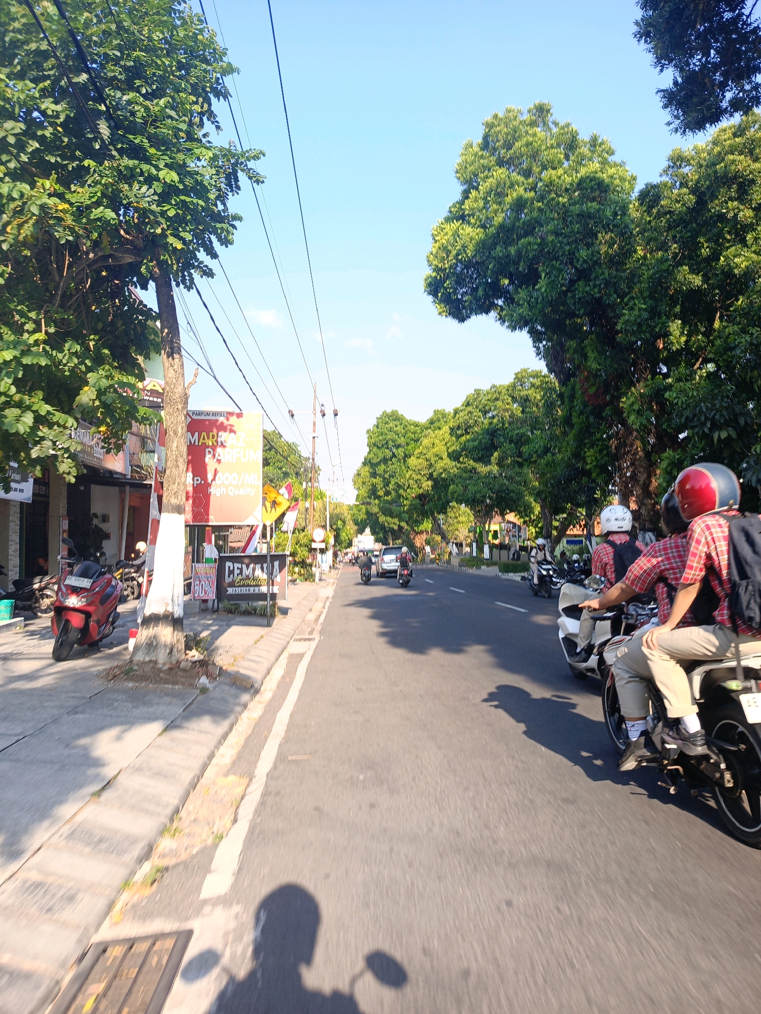 The image depicts a street scene in Candirejo, Indonesia, on a bright, sunny day. The viewpoint appears to be from a moving vehicle, likely a motorcycle, looking forward along a paved road. On the left side of the road, there are buildings with shops, trees, and utility poles with wires overhead. One shop has a large sign advertising "MARRAZ PARFUM" with prices. A red scooter is parked near the sidewalk. On the right side of the road, more trees line the street, casting shadows. Several motorcycles are present, with people riding them. In the foreground, two individuals are riding a motorcycle; the rider wears a red and blue helmet and a red and white plaid shirt with a backpack, while the passenger, wearing a white helmet, is visible from the back. The road is in good condition with visible lane markings. The overall environment suggests a typical urban or semi-urban setting in Indonesia.