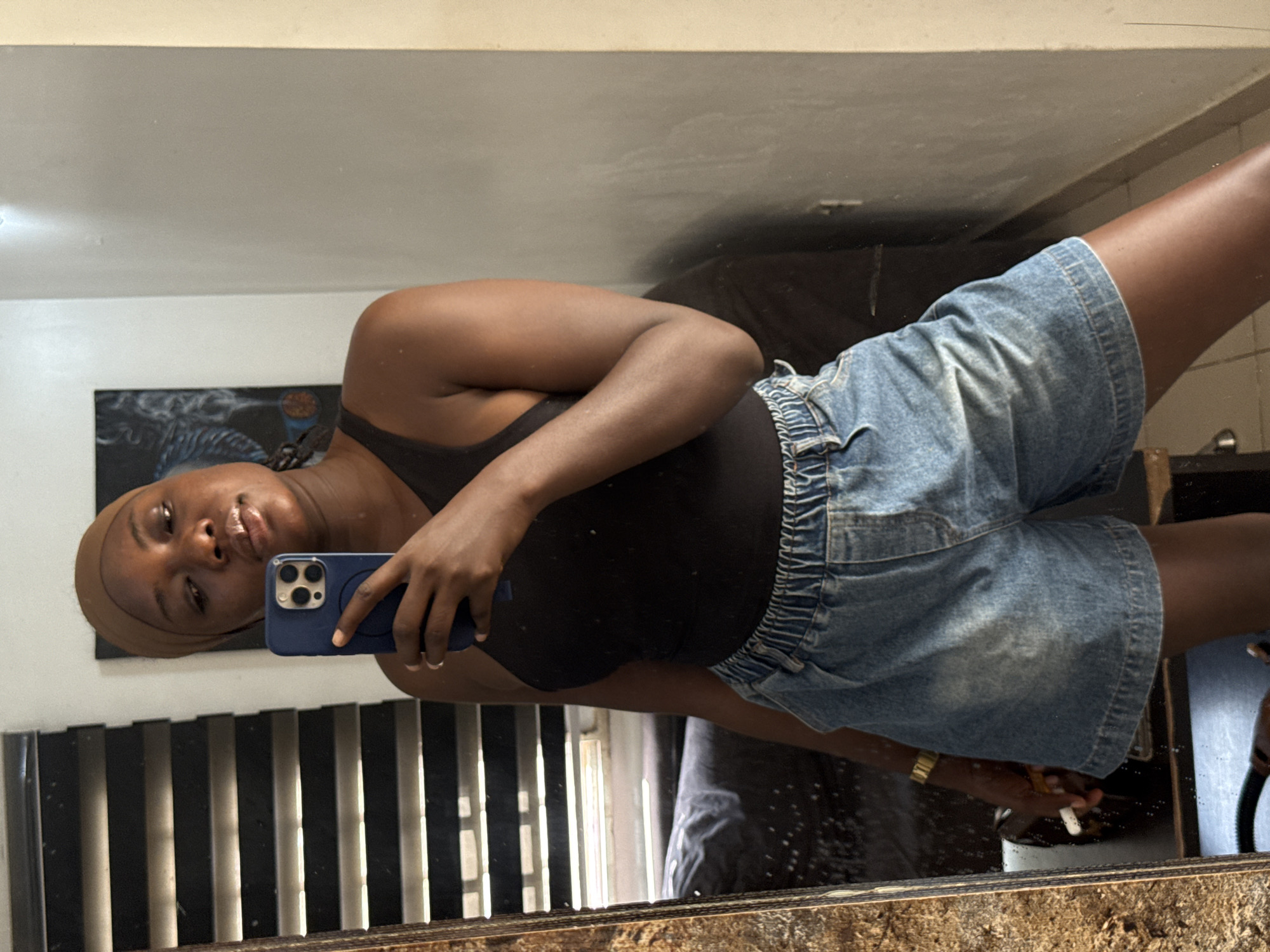 The media file displays a mirror selfie of a woman, oriented on its side, with the woman's head facing left. She is captured from the chest up to just above her knees, wearing a black tank top and light blue denim shorts with an elasticated waist. Her head is covered with a light brown cap or scarf, and she gazes directly at the camera with a neutral, slightly pensive expression, her lips slightly parted.
She holds a blue smartphone, identifiable as an iPhone with a three-lens camera system, in her left hand, positioned to capture her reflection in the mirror. A gold-toned watch adorns her right wrist. In her right hand, she holds a white cylindrical object, possibly a cigarette or a vaping device, with a subtle wisp of smoke or vapor visible. Her nails are short and natural.
The background, seen through the mirror, features a light-colored (off-white/cream) wall at the top, above a dark-toned abstract painting or artwork mounted on the wall. Below this artwork, a set of vertical black and white slats or blinds is visible, possibly from a window or furniture. The mirror rests on a dark, textured surface in the foreground, which appears to be wood. The indoor setting suggests a private room, likely in a home. The location context for this image is Abuja, Nigeria.