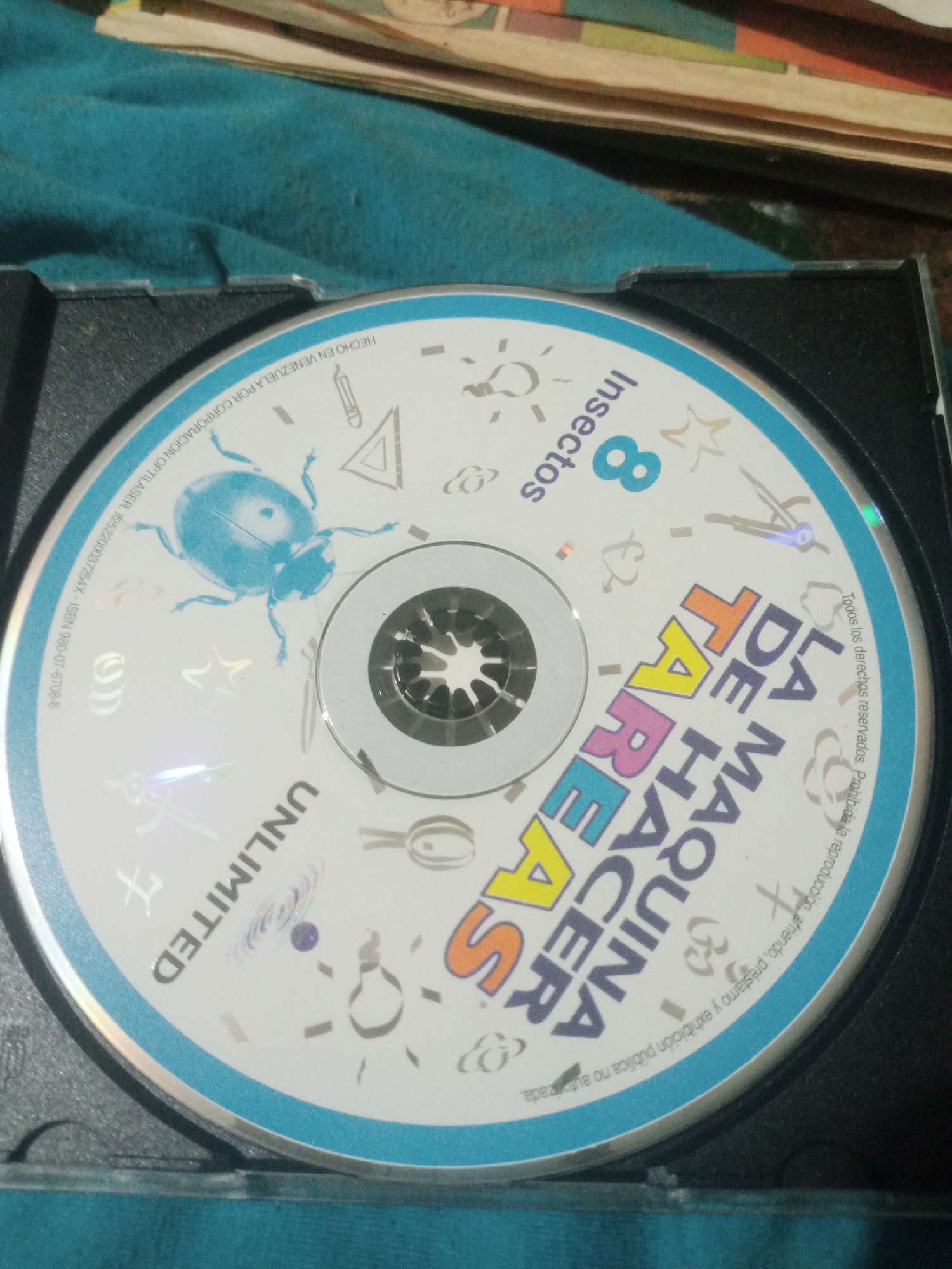 A CD is lying in a black CD case on a turquoise fabric surface. The CD's label is mostly visible and features a large, stylized blue ladybug graphic. Text on the CD includes "8 Insectos," "LA MAQUINA DE HACER TAREAS," and "UNLIMITED." There are also various smaller graphics of insects and geometric shapes scattered across the label. Along the outer edge of the CD label, text in a smaller font reads "HECHO EN VENEZUELA POR CORPORACION OPTILASER. I1E2S200037254X - ISBN 980-07-6708-8. Todos los derechos reservados. Prohibida la reproduccion, préstamo y exhibición pública no autorizada." The CD case is slightly ajar, revealing the reflective surface of the CD. The background consists of stacked books or papers. The scene is indoors. There is no discernible connection to the city of Santa Ana, Venezuela, beyond the text on the CD itself.