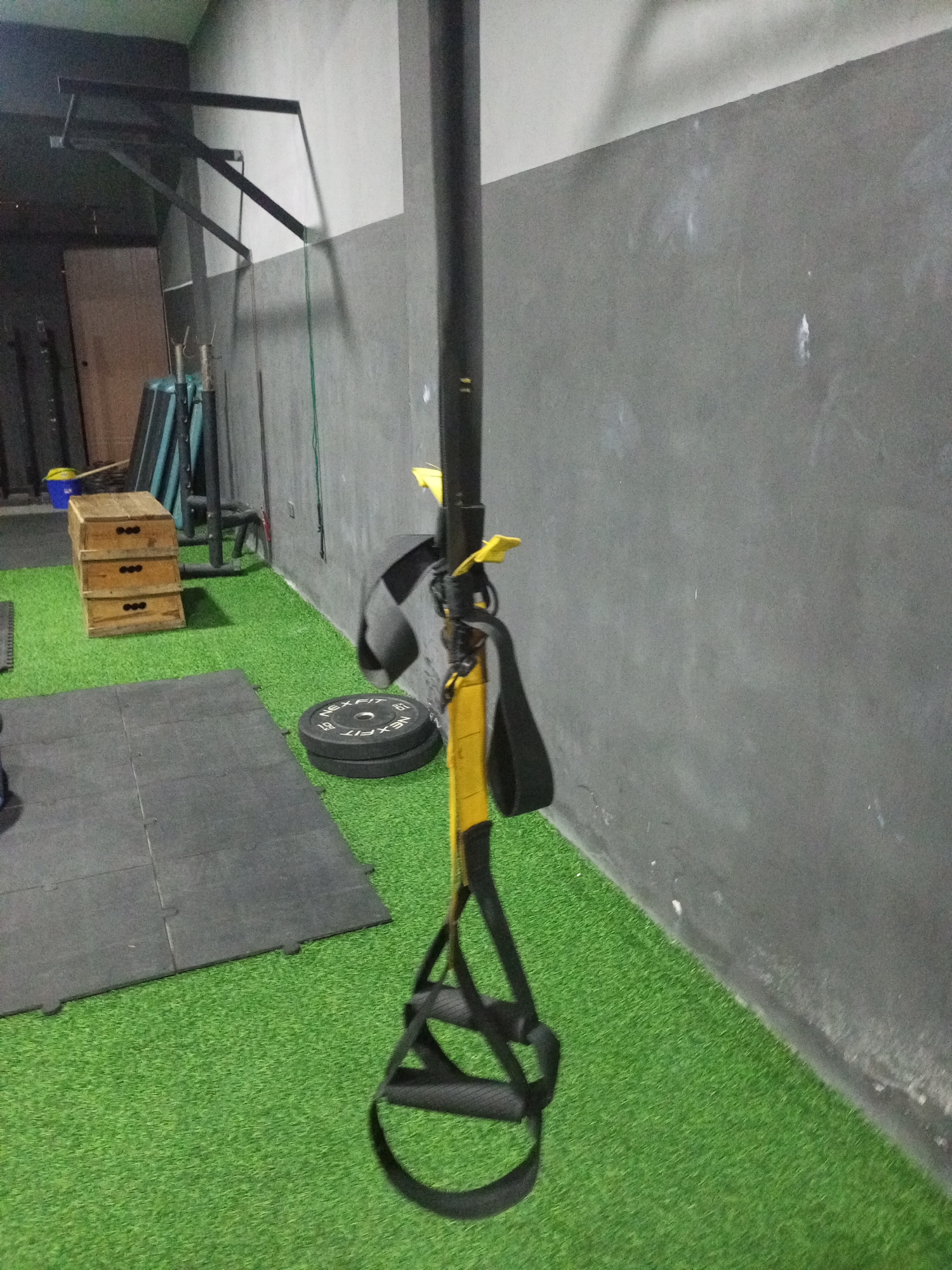 The image depicts an indoor gym with artificial turf flooring. In the foreground, a yellow and black suspension trainer hangs from a black pole. To the left, a stack of three wooden plyo boxes sits on the turf, with a blue bucket and a broom partially visible behind them. Next to the plyo boxes is a black rubber mat, and behind that, a stack of two weight plates. The background features a gray wall. In the upper left corner, a metal pull-up bar is mounted to the wall. The scene is static, with no people present. The lighting is artificial, casting shadows on the wall and floor. The overall impression is of a functional, albeit somewhat cluttered, workout space in Juan Griego, Venezuela.