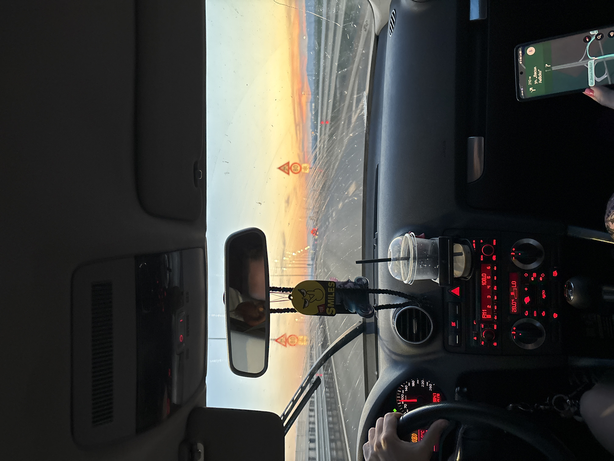 The image shows the interior of a car driving on a highway at sunset. The driver's hand is on the steering wheel, and the dashboard displays a speed of 70 km/h and a temperature of 28°C. A cup holder with a drink is visible, as well as a phone displaying navigation. The windshield has water droplets and streaks, suggesting recent rain. The road is a multi-lane highway with a concrete barrier. In the distance, traffic lights and signs are visible. The sky is filled with orange and yellow hues of the sunset. There is no explicit indication of the location being Mramor, Bulgaria, but the context of a highway and driving at sunset is clear.