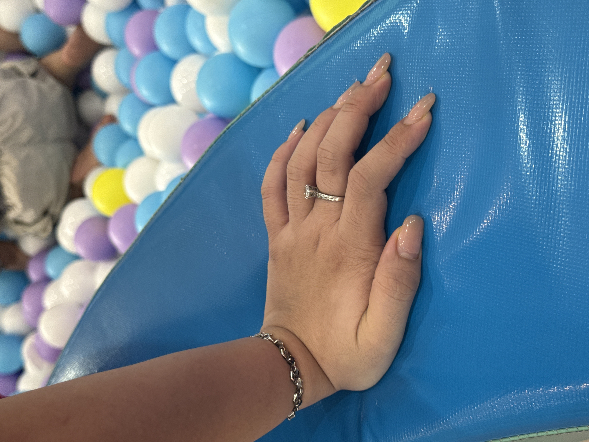 A person's hand, adorned with a silver bracelet and a ring with a small diamond, rests on a textured blue surface. In the background, a collection of colorful balls in shades of white, purple, and blue are visible. The setting appears to be a ball pit. The image was taken in Bac Nha Trang Ward, Vietnam.