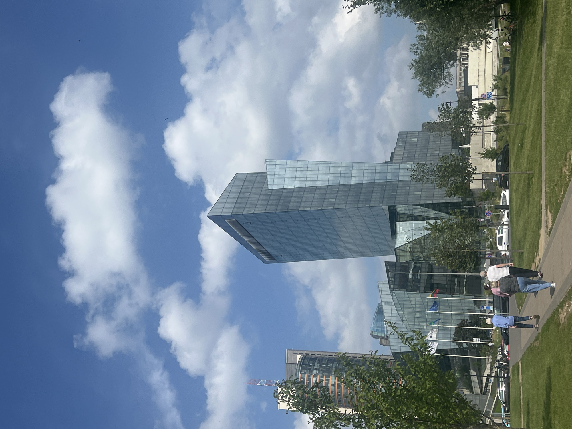 This is an exterior, eye-level, vertical, full-shot photograph taken on a bright, sunny day with scattered clouds. The focal point is a modern, glass-facade skyscraper with angular geometric designs. In the foreground, a paved walkway curves through a manicured green lawn. Four people, two couples, walk away from the camera on the walkway. To the left, a cluster of trees partially obscures the view of another skyscraper with a red and white antenna. To the right of the main skyscraper, there are other glass-facade buildings and trees. The scene is in the business district of Vilnius, Lithuania.