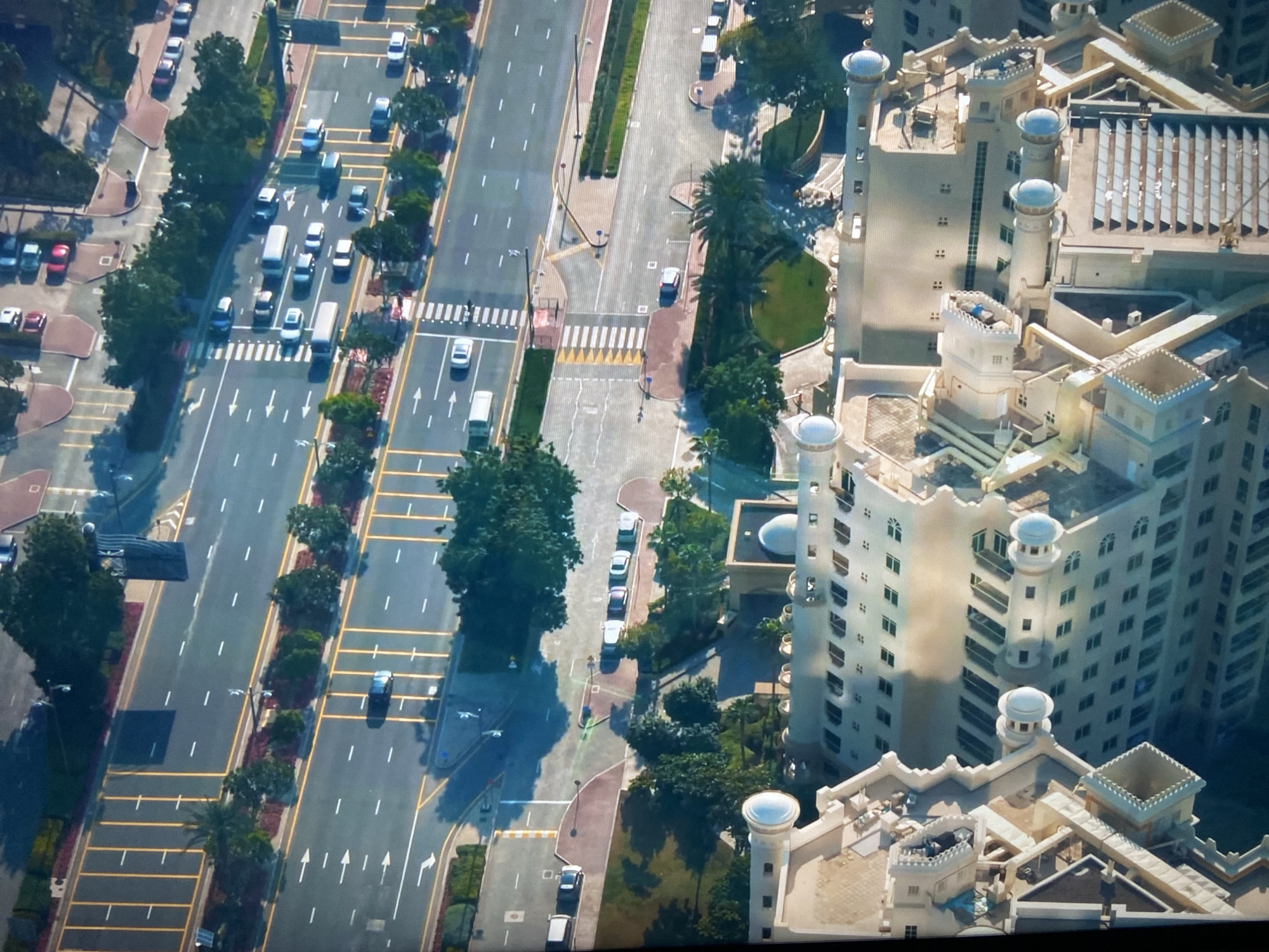 An aerial photograph shows a section of a city with a straight multi-lane road running parallel to a tall, light-colored building complex. The road is divided into several lanes with white lane markings and is dotted with vehicular traffic moving in both directions. Pedestrian crossings with yellow and white stripes are visible at intervals along the road. Trees and other greenery line the roadsides. Adjacent to the road, on the right side of the image, is a sidewalk with parked cars. The building complex has a light facade with a distinctive architectural style, featuring towers with rounded tops and intricate patterns. The architecture of the building suggests it could be a luxury hotel or residential complex. The image is taken from a high angle, providing a comprehensive view of the urban landscape. The location appears to be a densely populated area, possibly a metropolitan city.