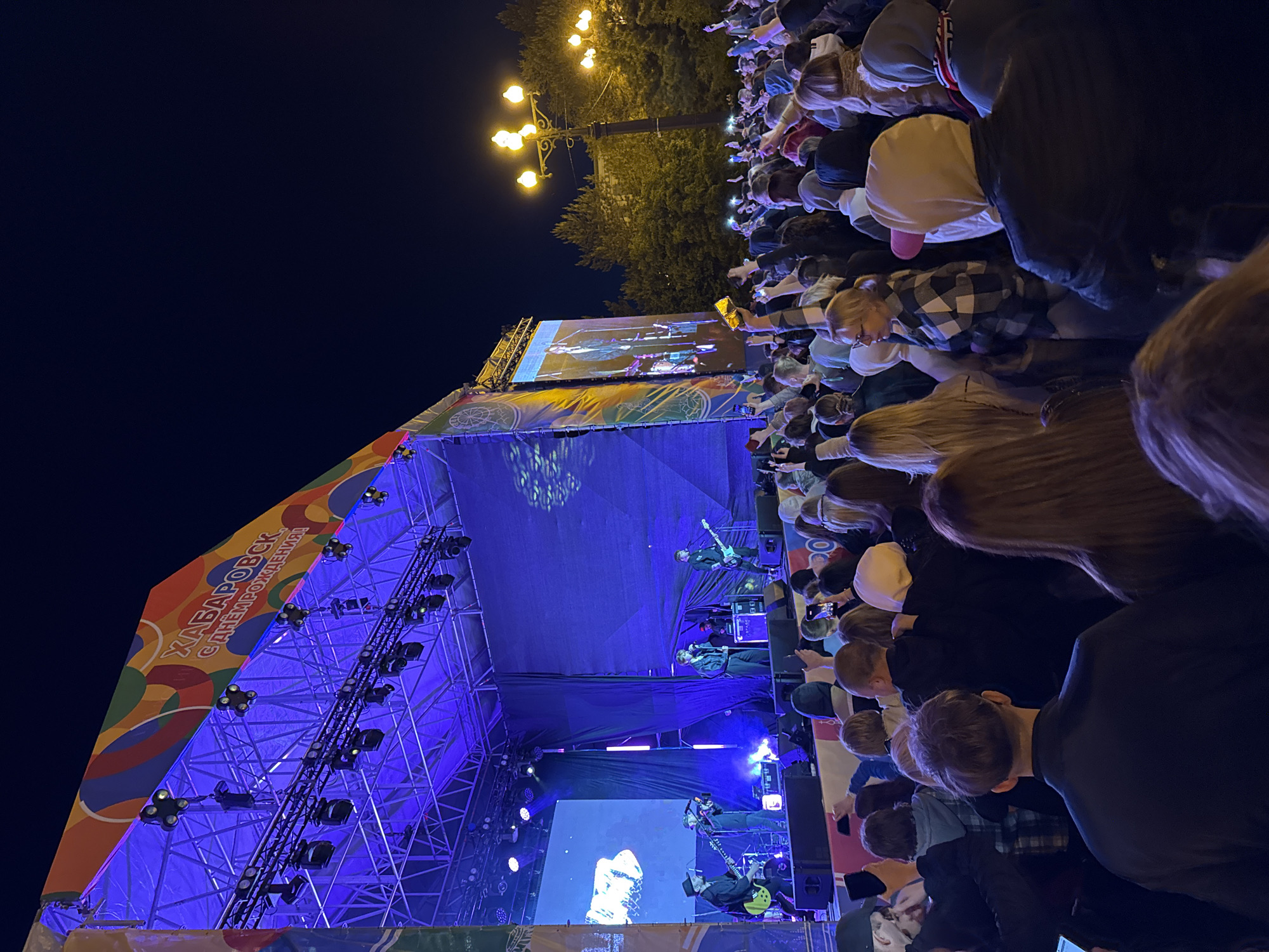 At night, a crowd watches a performance on a large outdoor stage in Khabarovsk, Russia. The stage is illuminated with blue lights and features large screens displaying images. The crowd is dense, consisting of people of varying ages and clothing. Some are holding up phones, presumably to record the event. In the background, a lamppost with multiple lights is visible. A banner is attached to the left of the stage and features the name of the city.