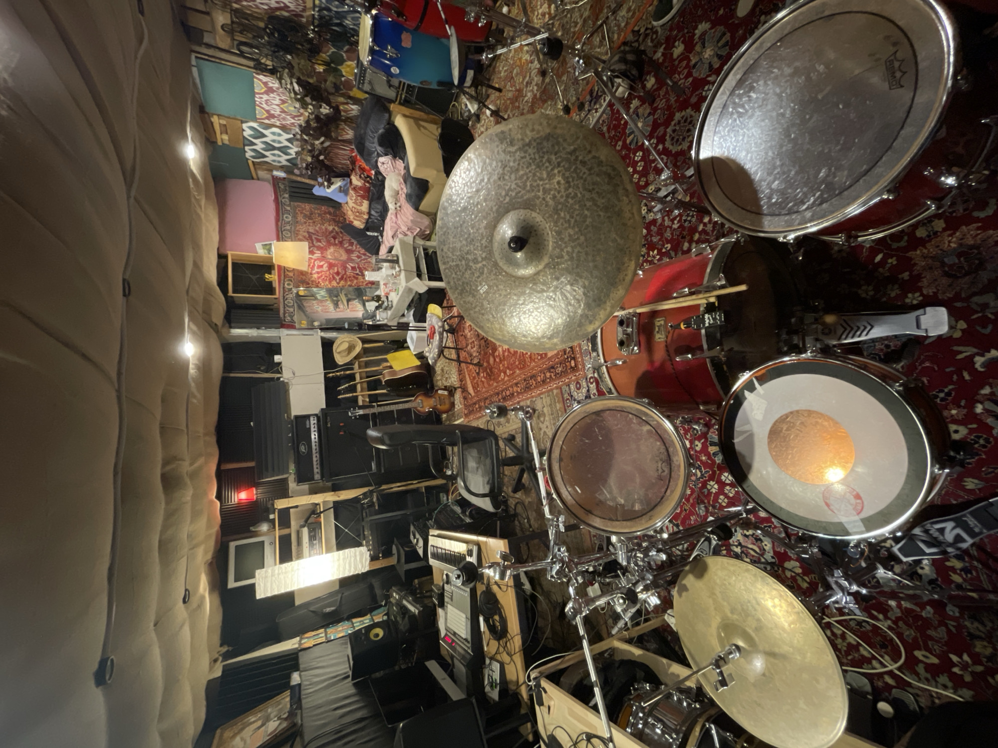 The image shows a drum set in a music studio. The drum set is red, with various cymbals. In the background, there is music equipment, including amplifiers and guitars. There are also rugs on the floor. The studio appears to be located in a room with soundproofing on the walls. There are no people visible in the frame. The location in the city of Yekaterinburg, Russia cannot be inferred from the image.