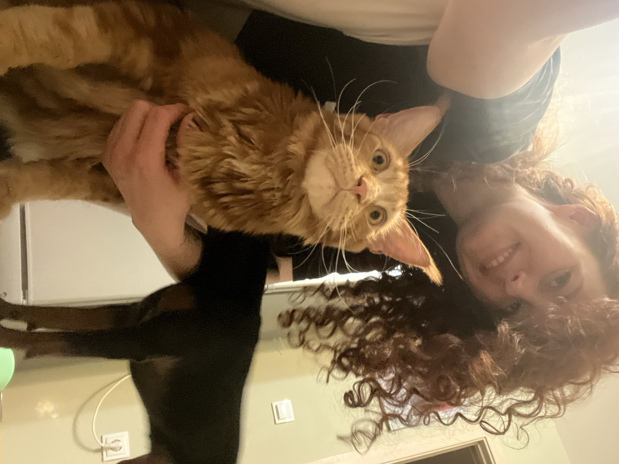 A woman with curly brown hair is holding a large, ginger Maine Coon cat. The woman is smiling at the camera. The cat is looking directly at the viewer with wide, yellow eyes. The background appears to be an interior space with a black object, possibly another animal, partially visible. A white electrical outlet and a light switch are visible on the wall. The location of the image in Saint Petersburg, Russia cannot be determined from the given image.